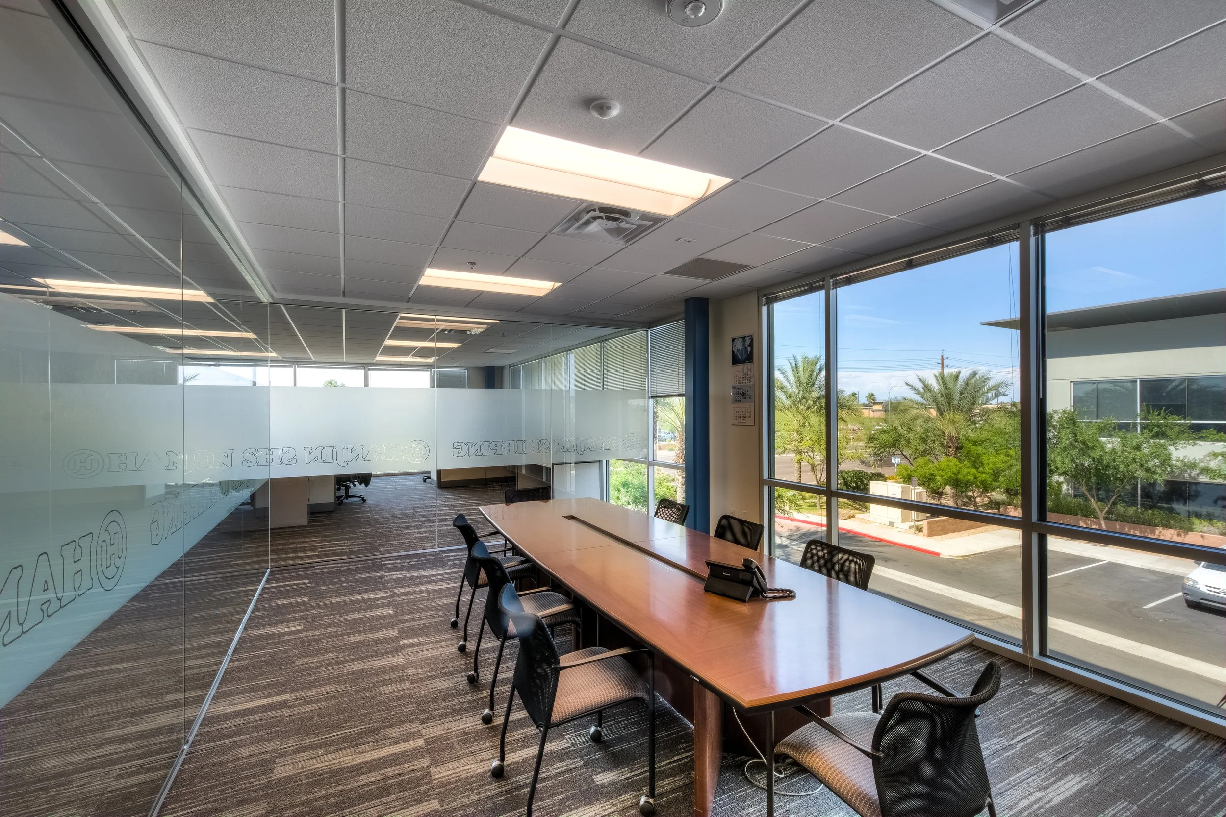 Empty conference room with large wooden table, black chairs, and a phone, with floor-to-ceiling windows showing trees and a building outside.
