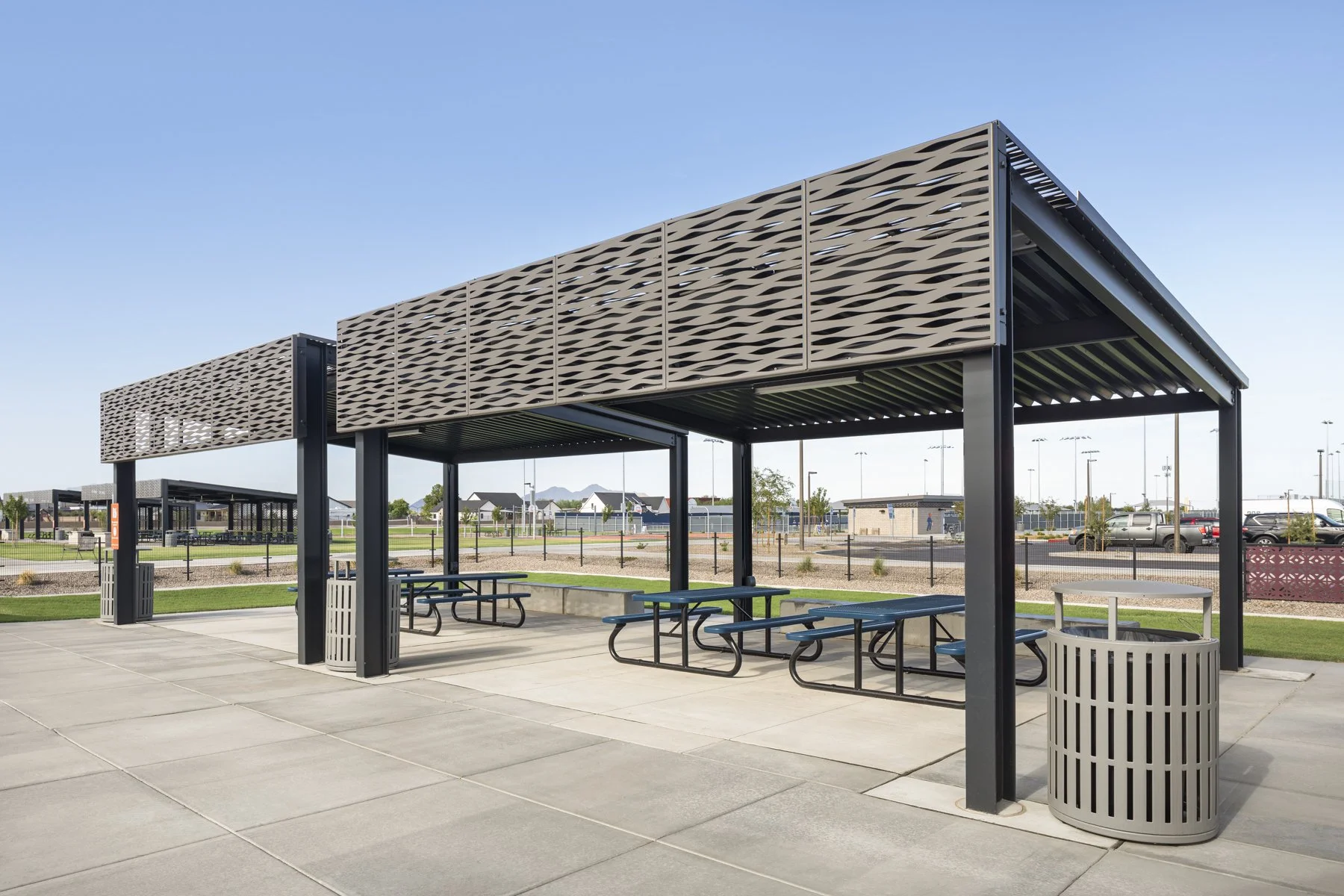 Empty outdoor picnic shelter with multiple blue picnic tables and a trash can, located in a park with a paved area and parking lot in the background under a clear blue sky.