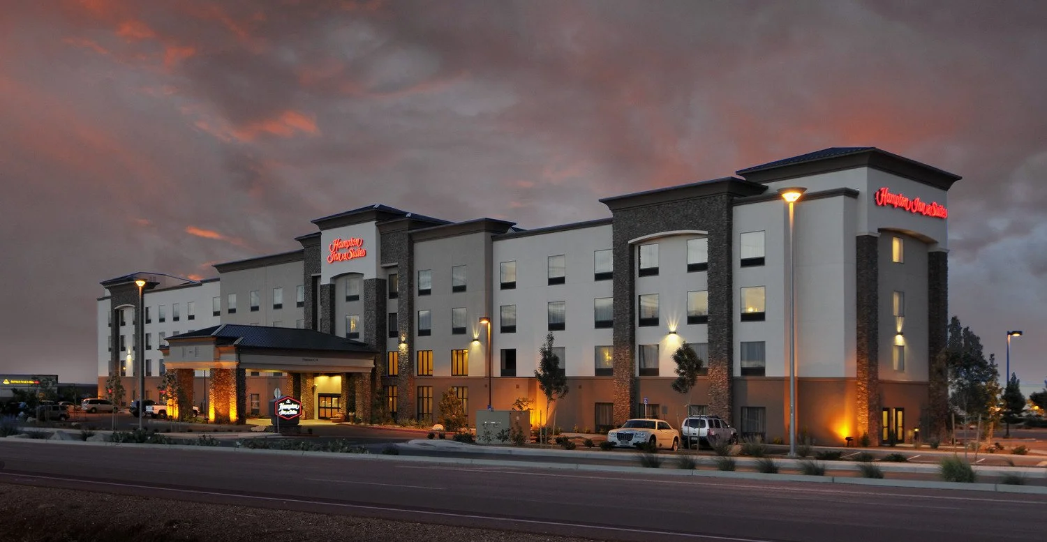 A modern Hilton Garden Inn hotel with white and gray exterior walls, lit up at dusk with outdoor lights, parked cars, and a dark, cloudy sky in the background.
