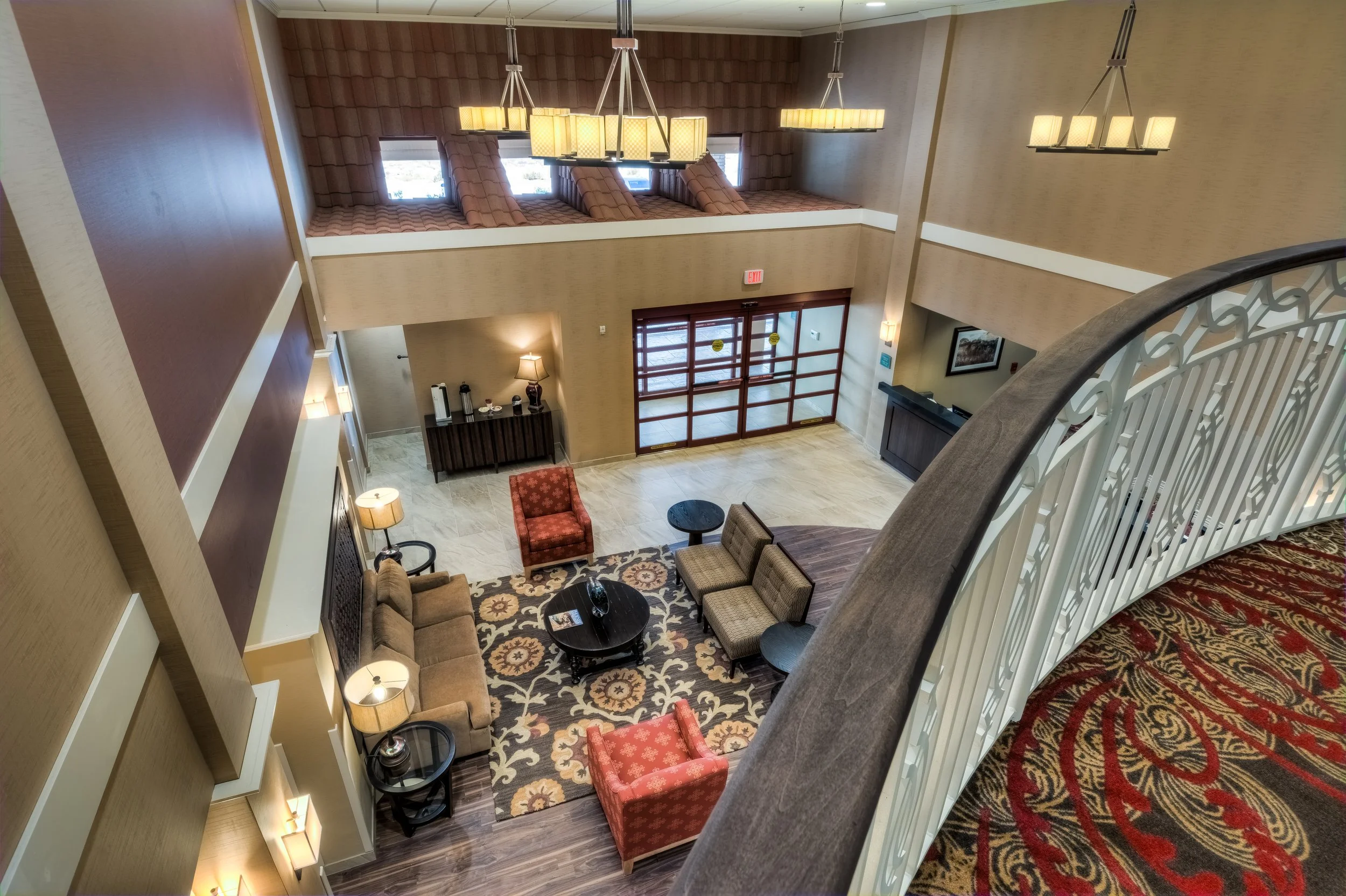 View of a hotel lobby from an upper balcony showing seating area with sofas, armchairs, coffee tables, a patterned rug, a side table with a lamp, and the entrance with glass doors, and decorative lighting fixtures hanging from the ceiling.