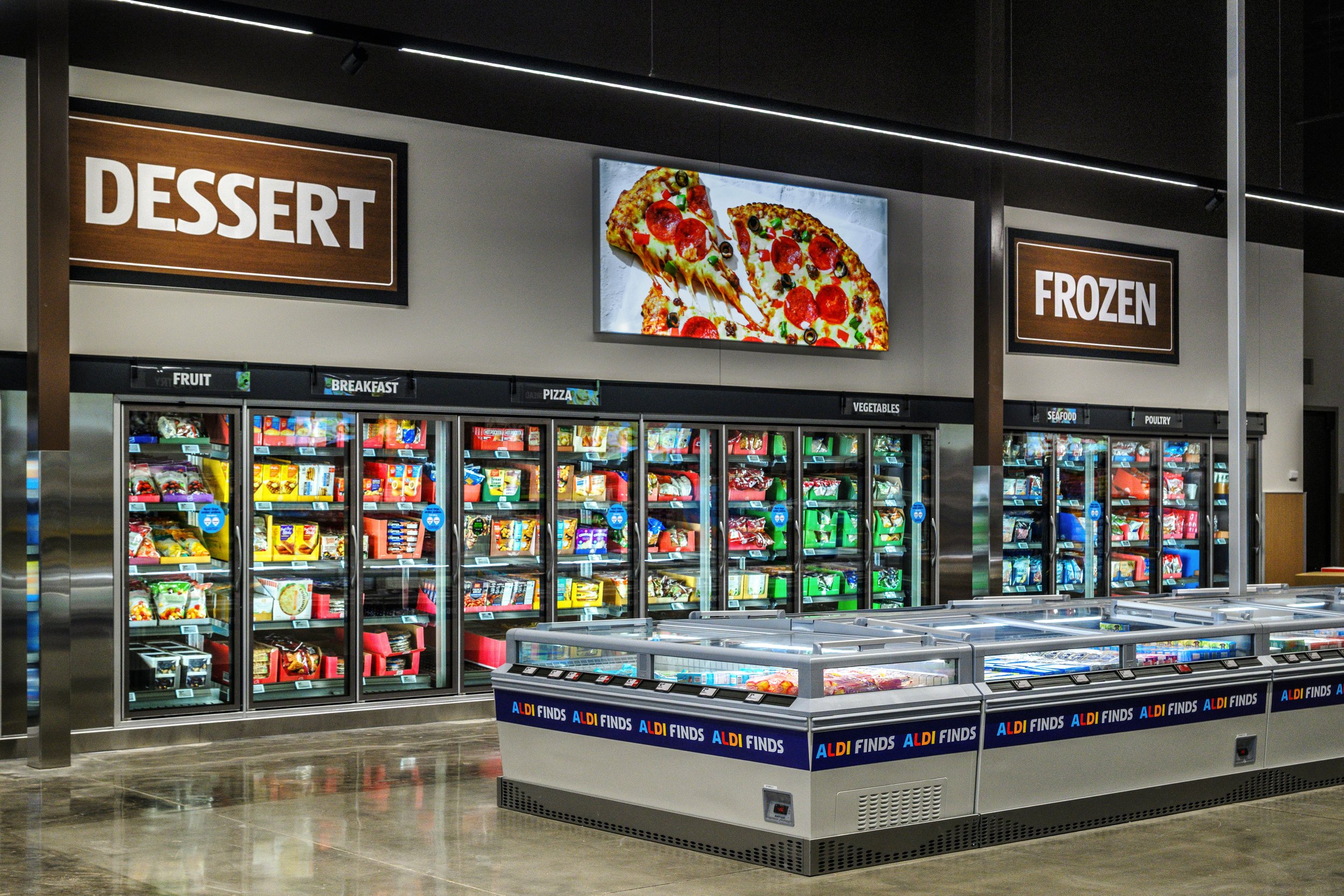 Grocery store aisle with frozen foods and refrigerated sections labeled 'DESSERT' and 'FROZEN' with digital screens displaying pizza images and various frozen items inside glass-freeze doors.