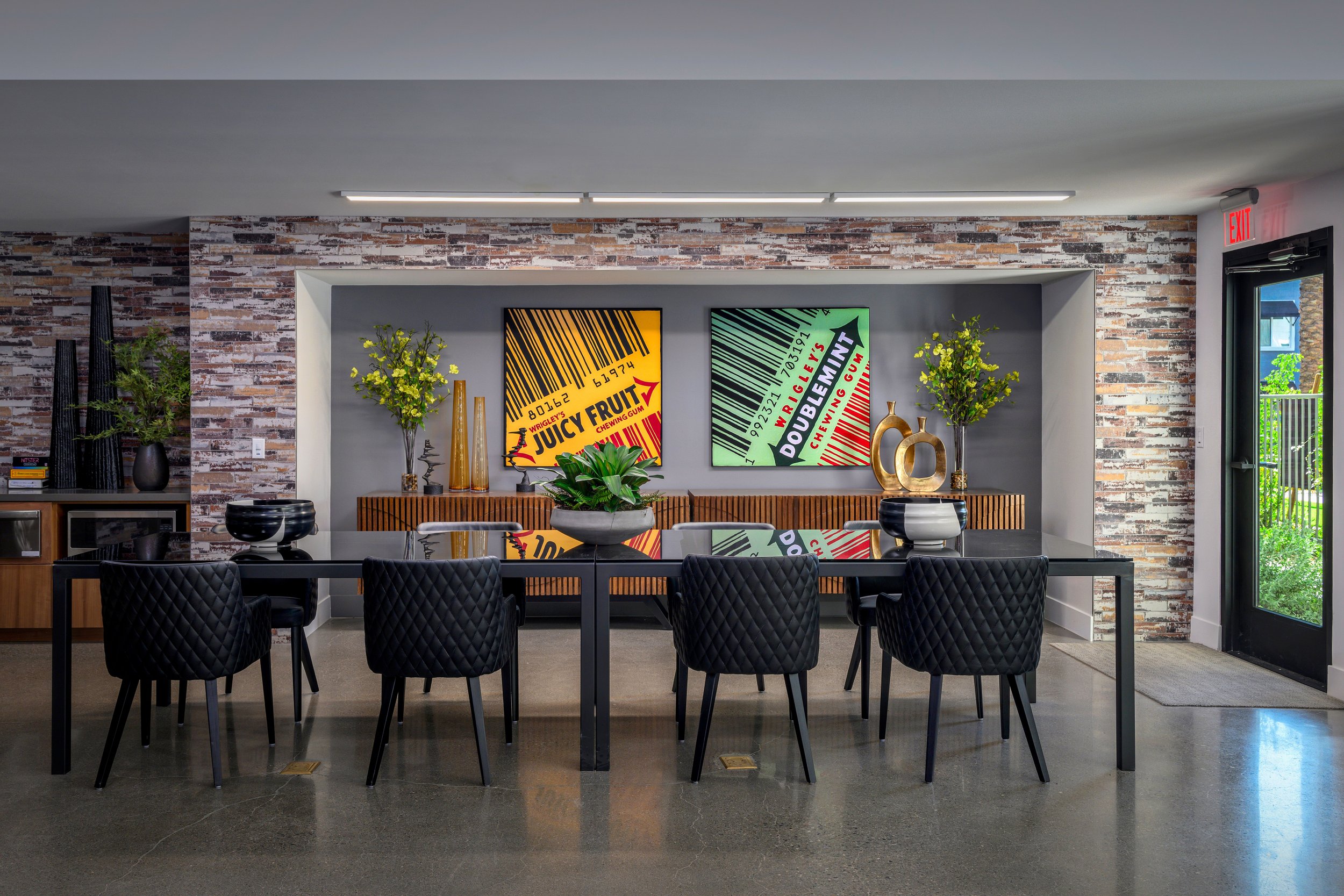 Modern dining room with a large black table, six black quilted chairs, a brick accent wall, two colorful pop art style prints of chewing gum packages, decorative vases, green plants, and a glass door to the outside with a garden view.