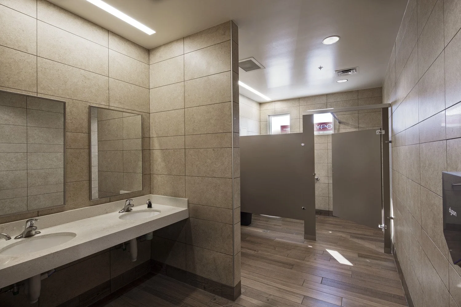 Public men's bathroom with two sinks, mirrors, and partitioned stalls, beige tiled walls and wood-patterned flooring.