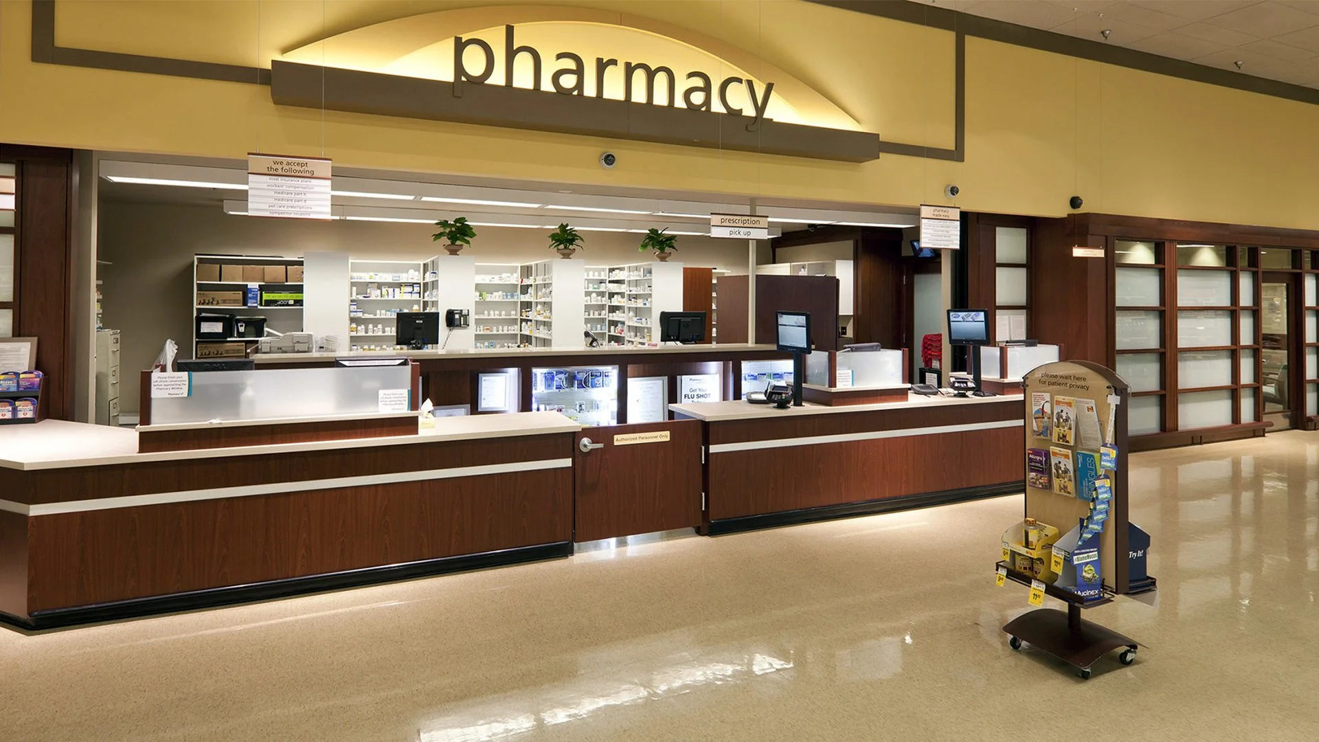 Empty pharmacy counter with shelves of medications behind, and a display stand with health products in the foreground.