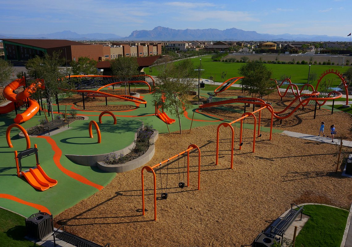 Children's playground with orange slides, swings, and climbing structures set on a green and brown surface, with residential neighborhood and mountains in the background.
