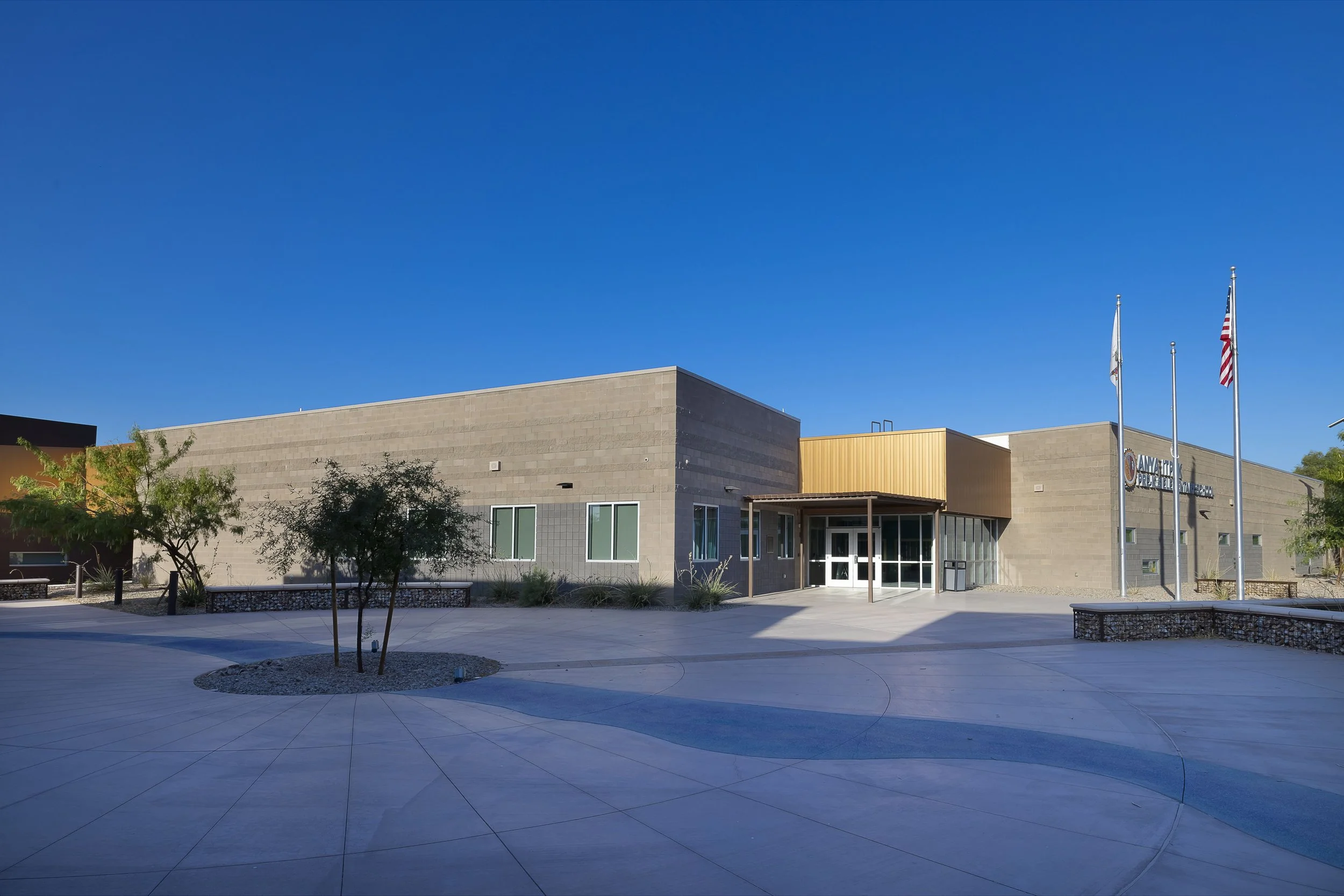 Exterior of a modern school building with a wide concrete courtyard, small trees, flags, and a clear blue sky.