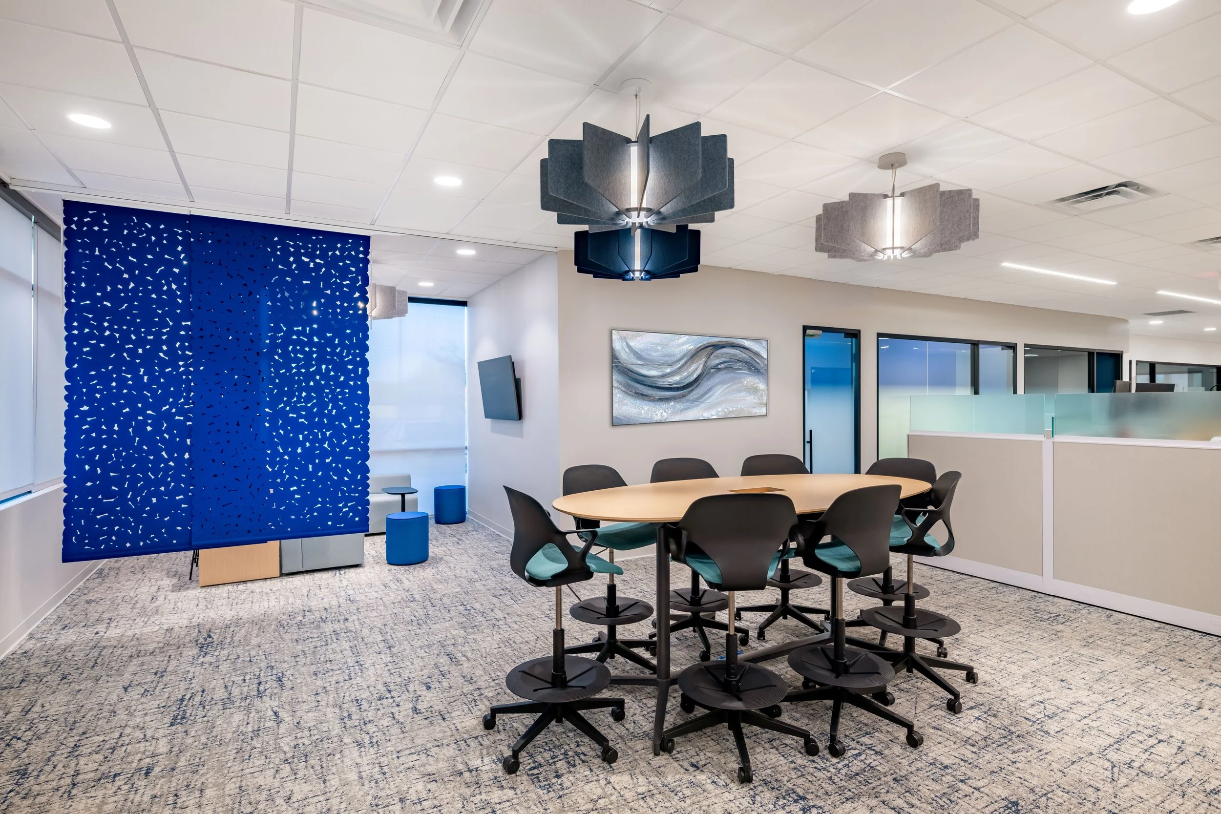 Modern office conference room with a round table, eight black and teal office chairs, abstract wall art, decorative ceiling lights, a blue partition, and floor-to-ceiling windows.