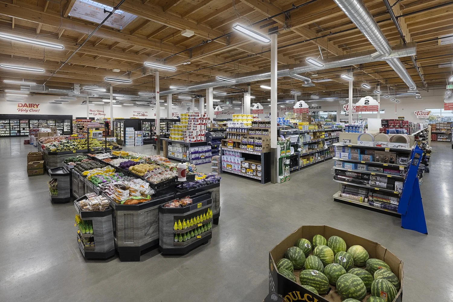 Interior of a grocery store with produce displays and aisles of packaged goods, exposed wooden ceiling, and ductwork.