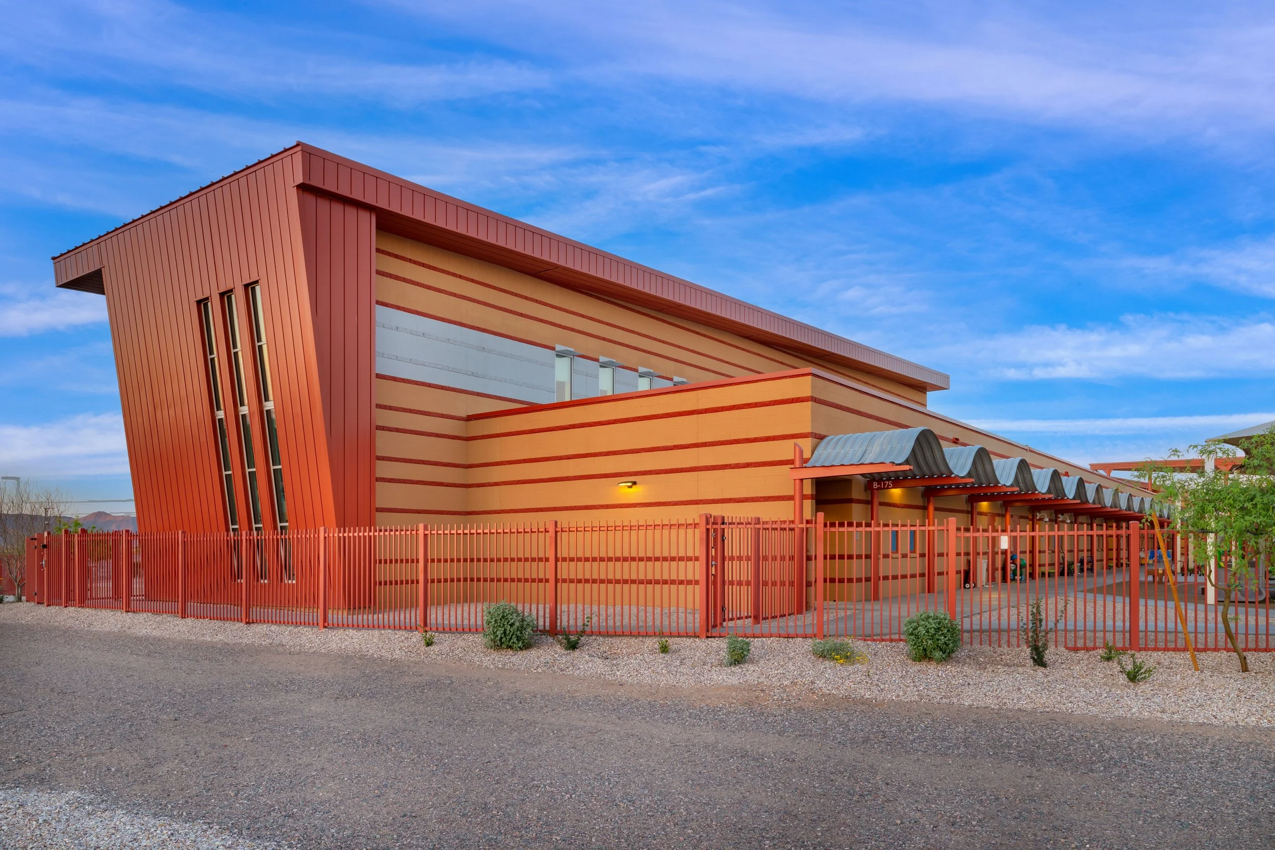 A modern building with orange and beige horizontal stripes, red metal fencing, and a curved metal awning over the entrance, under a blue sky with wispy clouds.