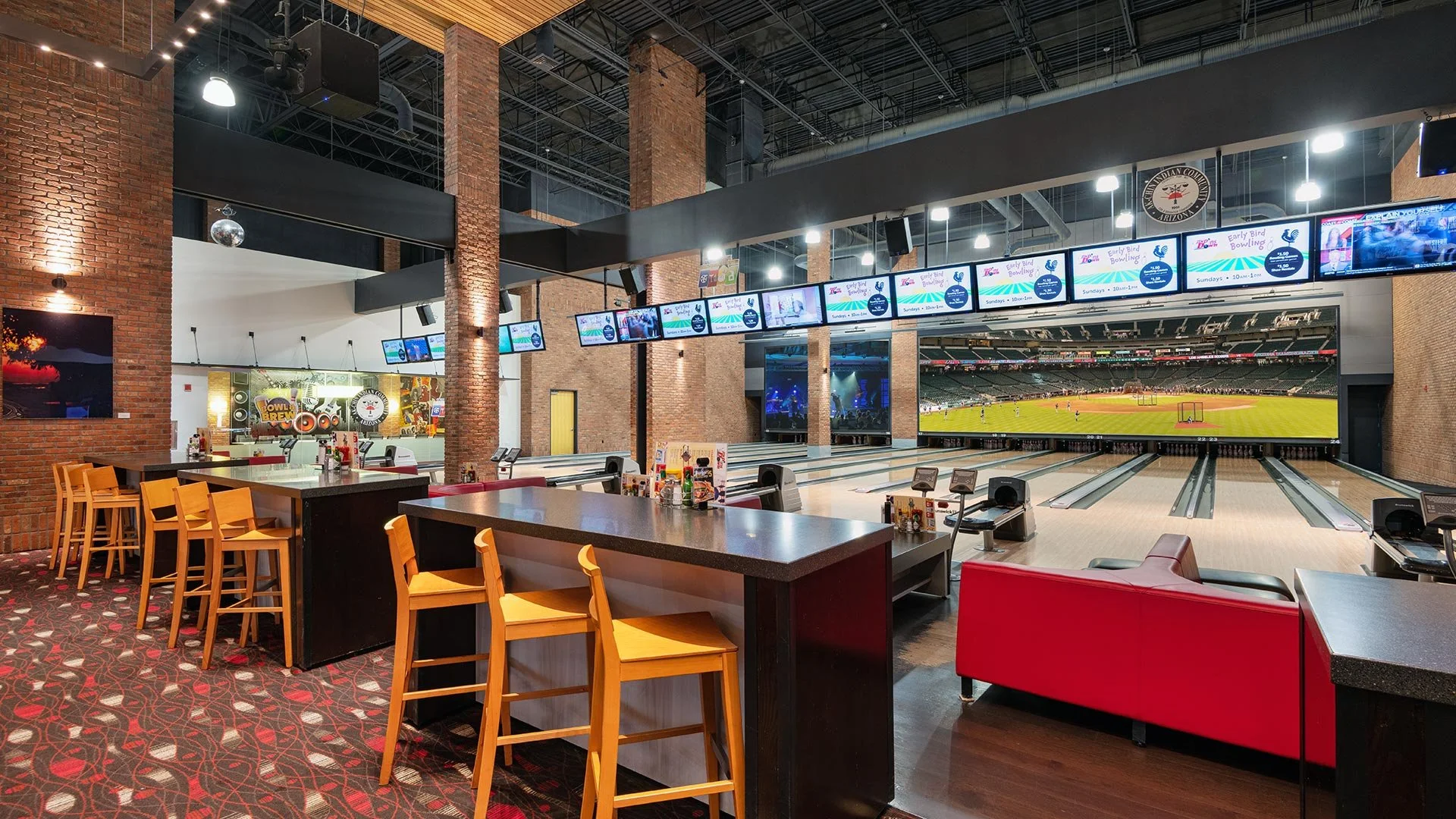 Interior of a bowling alley with a large playing area, multiple bowling lanes, electronic score screens, a bar with high chairs, and a large screen displaying a baseball game.