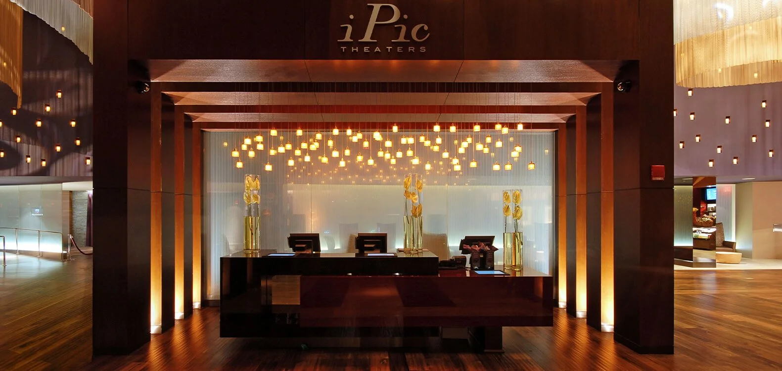 Hotel reception desk at iPic Theaters, featuring a dark wooden counter, illuminated hanging lights, and two tall vases with white orchids.