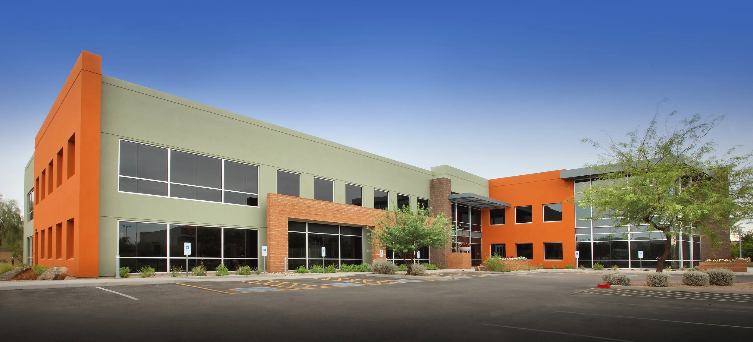 Modern commercial building with green and orange exterior walls, large windows, and a parking lot with designated handicapped parking spots, surrounded by desert landscaping.