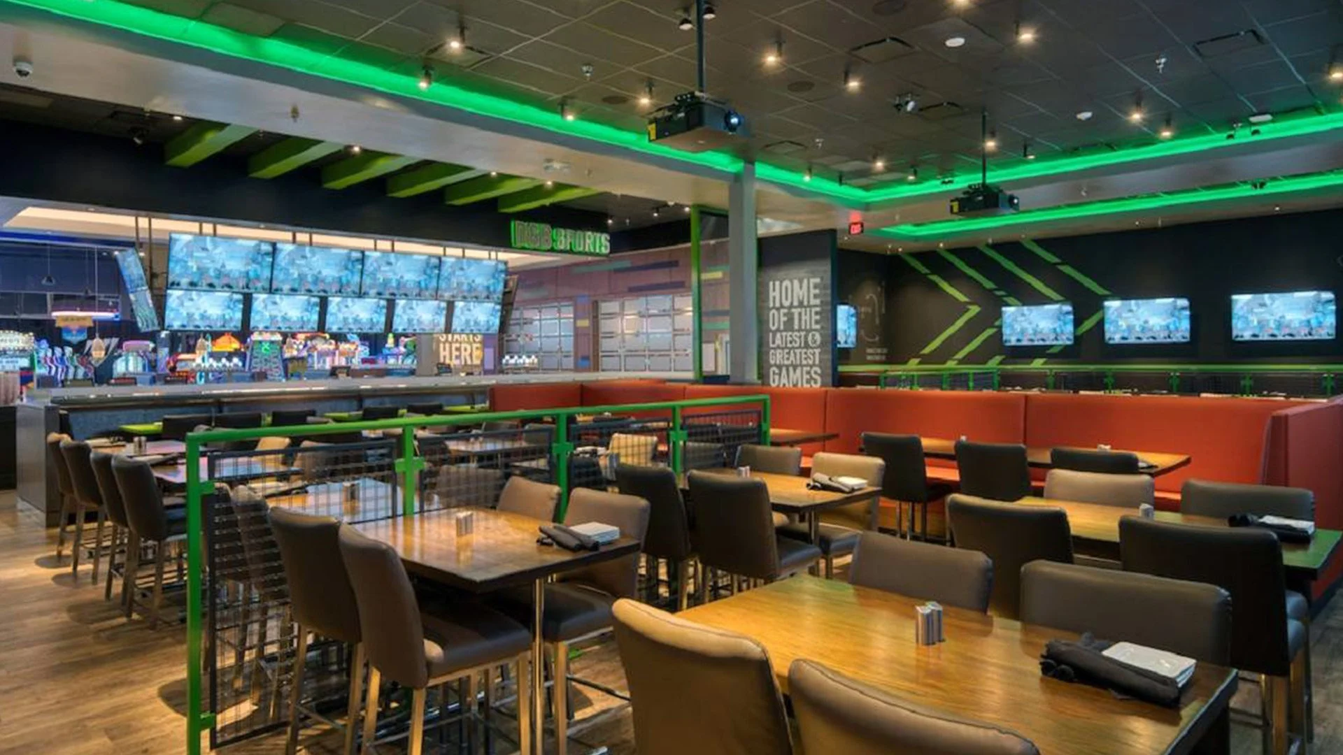 Interior of a sports bar with multiple tables and chairs, large TV screens displaying sports, neon green and black decor, and a bar area in the background.