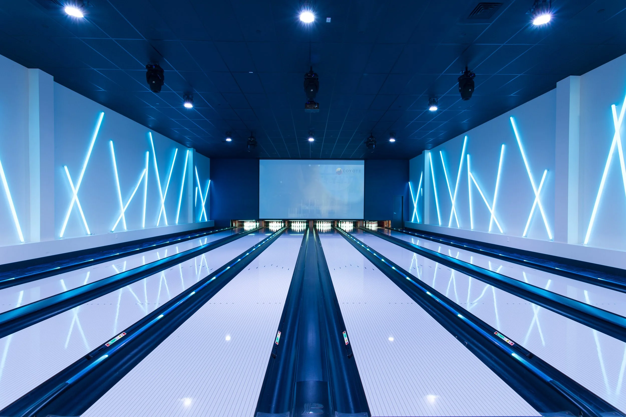 Modern bowling alley with four lanes, illuminated by blue LED strip lights on the walls, a large screen at the end of the lanes, and black ceiling with ceiling lights.