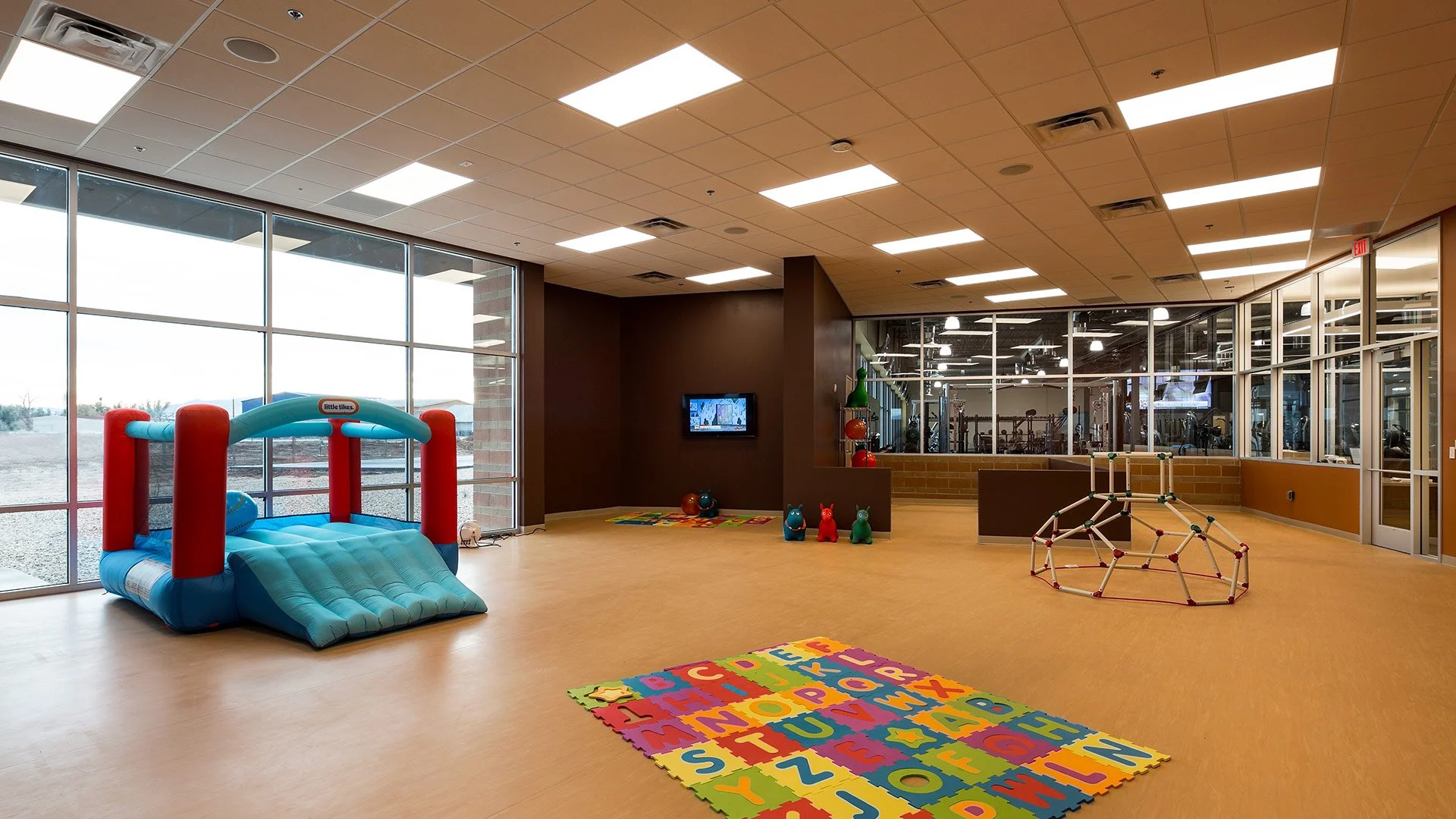 Indoor children's play area with a blue and red bouncy house, colorful alphabet foam mat, and a small climbing structure. The room has large windows, wooden flooring, and a view into a gym.