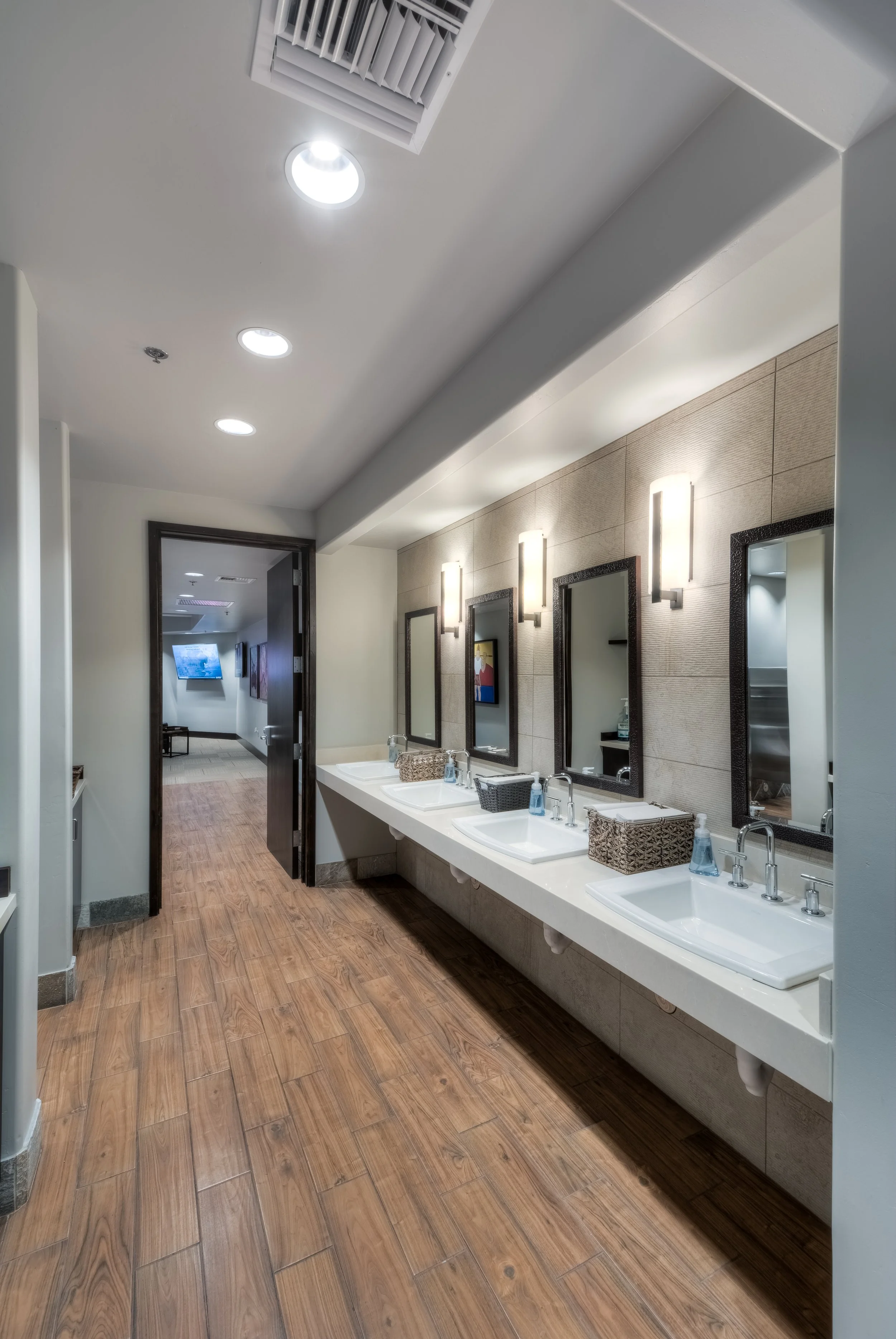 Public restroom with four sinks, mirrors, and wall-mounted lights, wood-look floor, hallway leading to another area with a television.