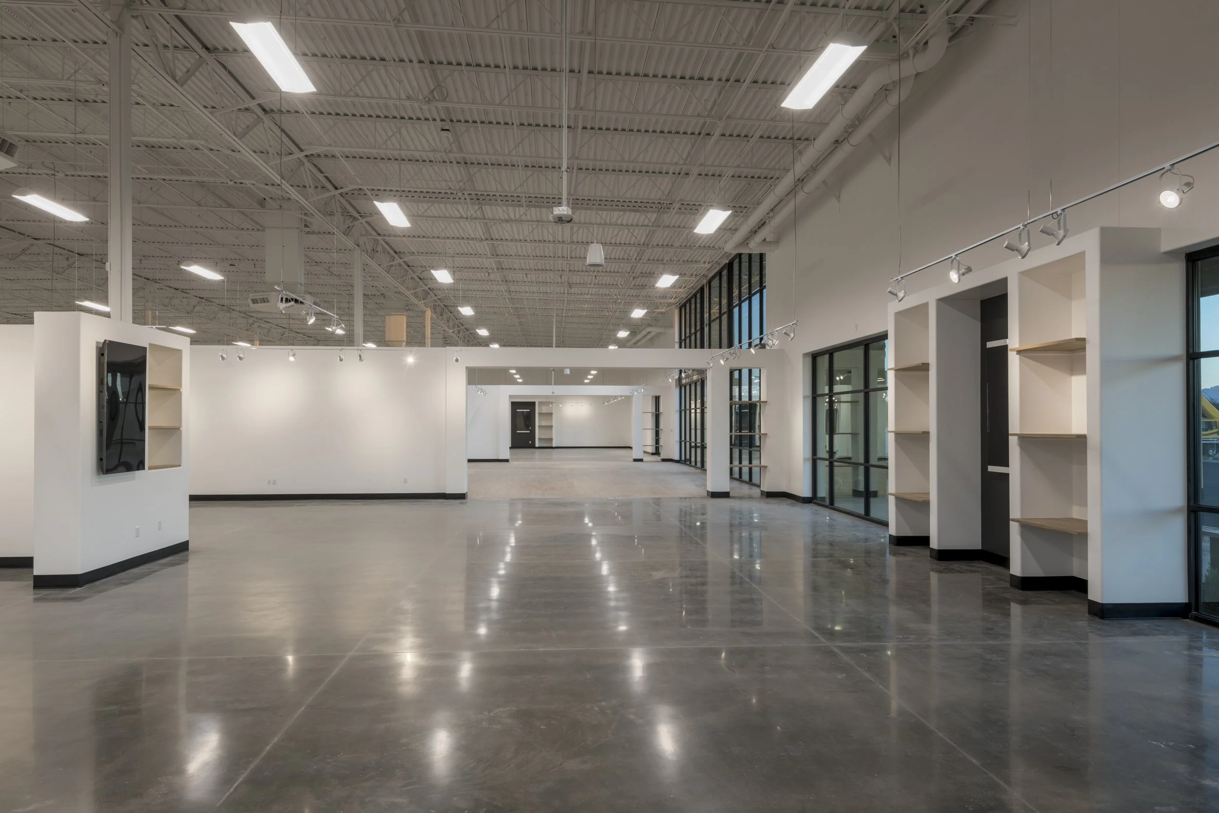 Interior of a large, empty retail or gallery space with polished concrete floors, white walls, ceiling track lighting, and large floor-to-ceiling windows.
