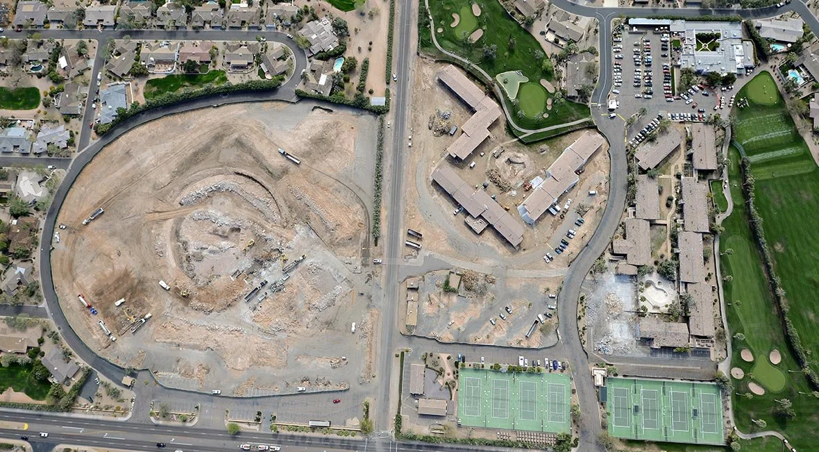 Aerial view of a construction site in a residential area, with some tennis courts and green fields nearby.