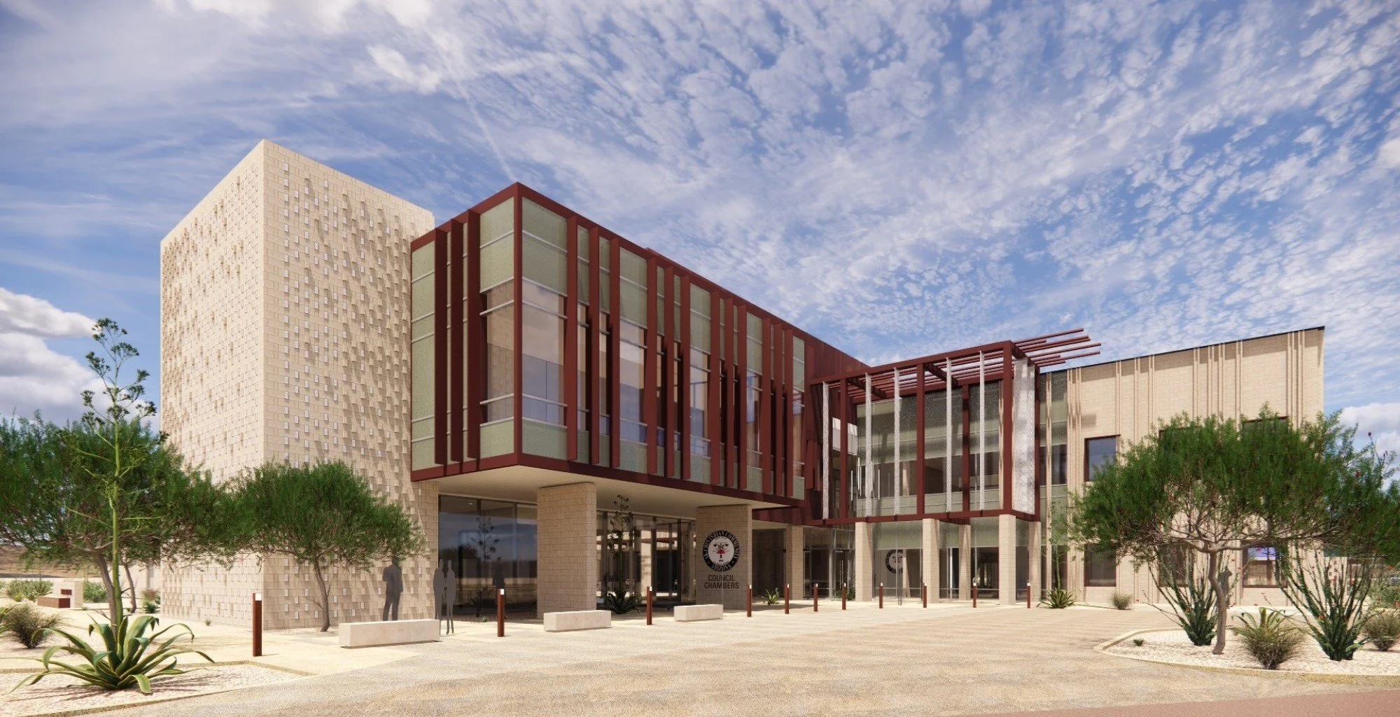 Modern city hall building with beige and red architectural design, surrounded by desert landscaping and small trees, under a partly cloudy sky.
