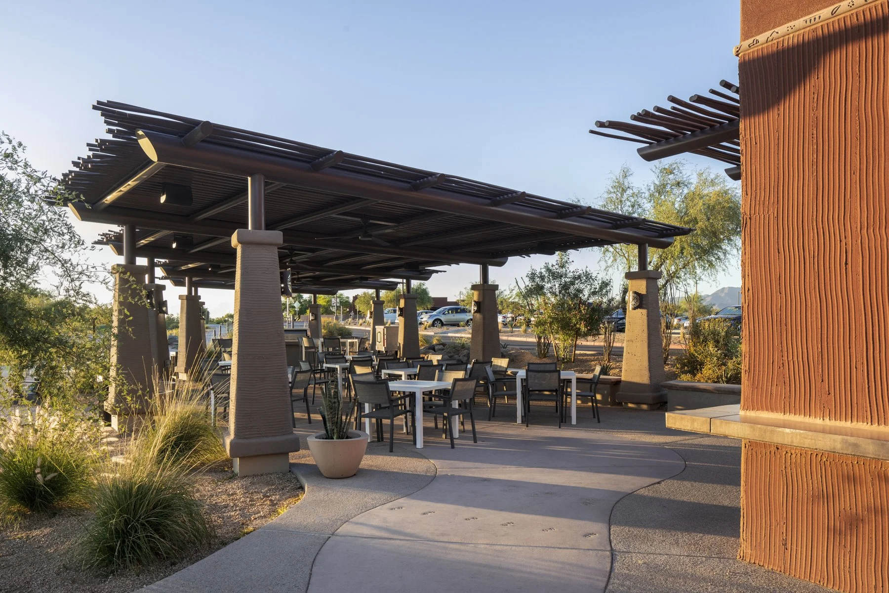 Outdoor patio with tables, chairs, and a shaded roof structure at sunset or late afternoon, surrounded by desert landscaping and mountains in the background.