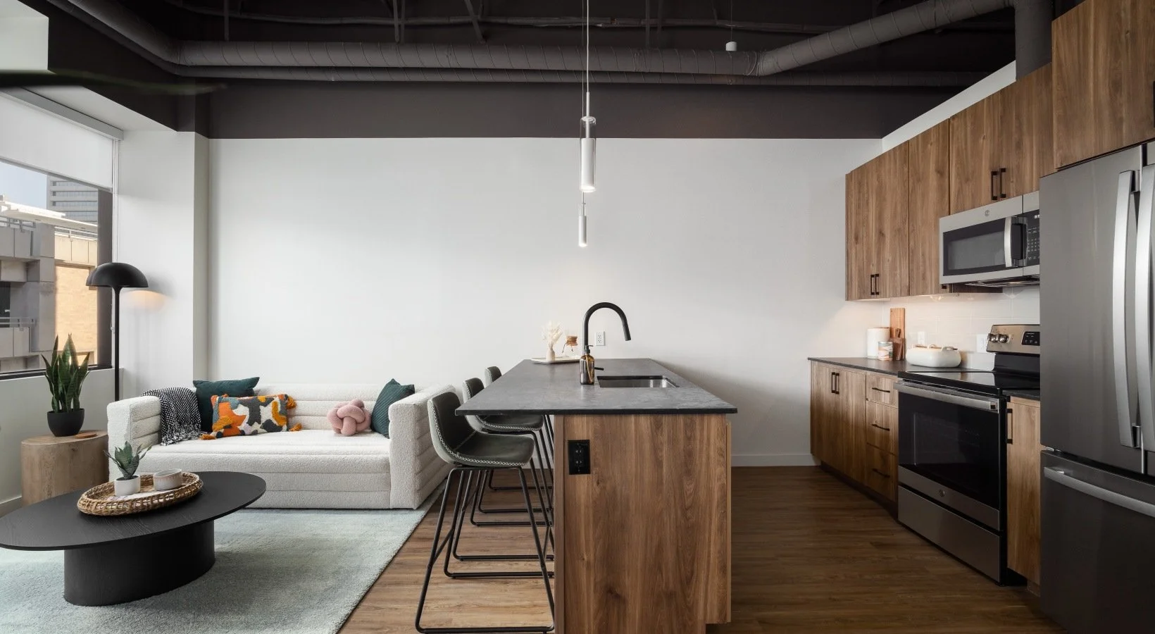 Modern open-concept kitchen and living room with a white couch, decorative pillows, a black coffee table, wooden flooring, and natural light from large windows.