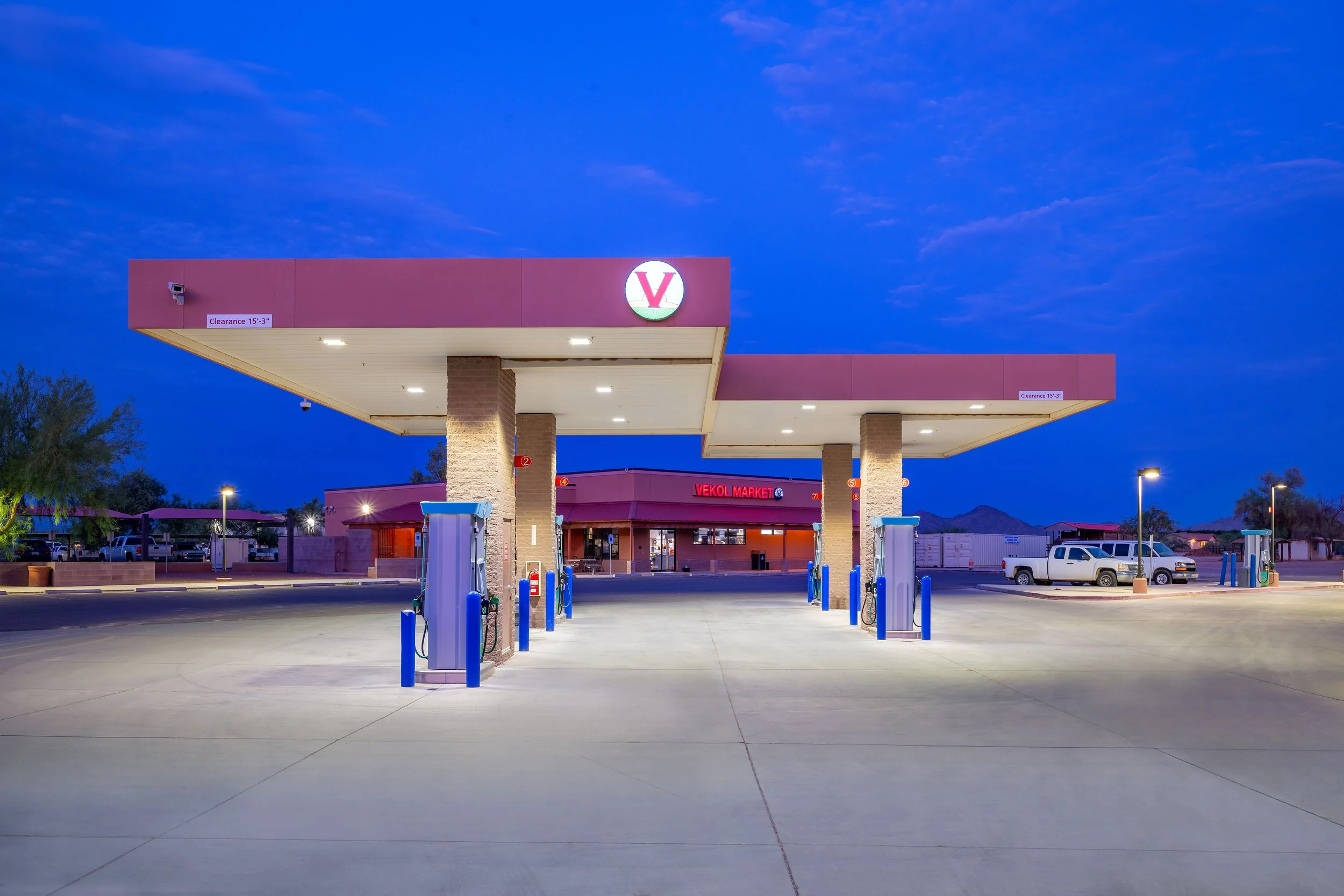 Empty gas station with multiple fuel pumps and a convenience store in the background during dusk, with mountains and trees visible in the distance.
