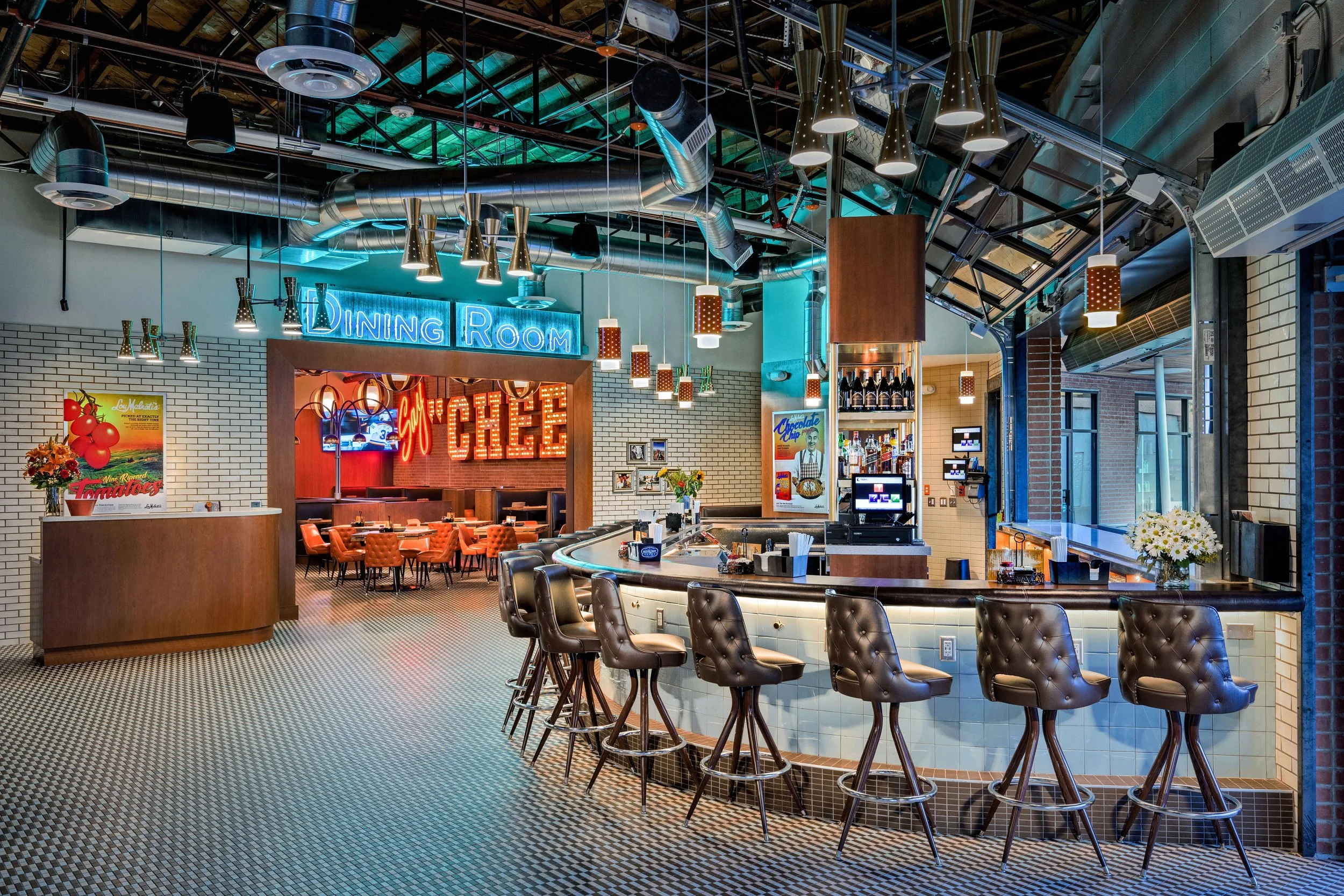 Interior of a modern restaurant with a bar in the foreground, barstools, a neon sign that says 'Dining Room,' and a dining area with tables and chairs in the background.