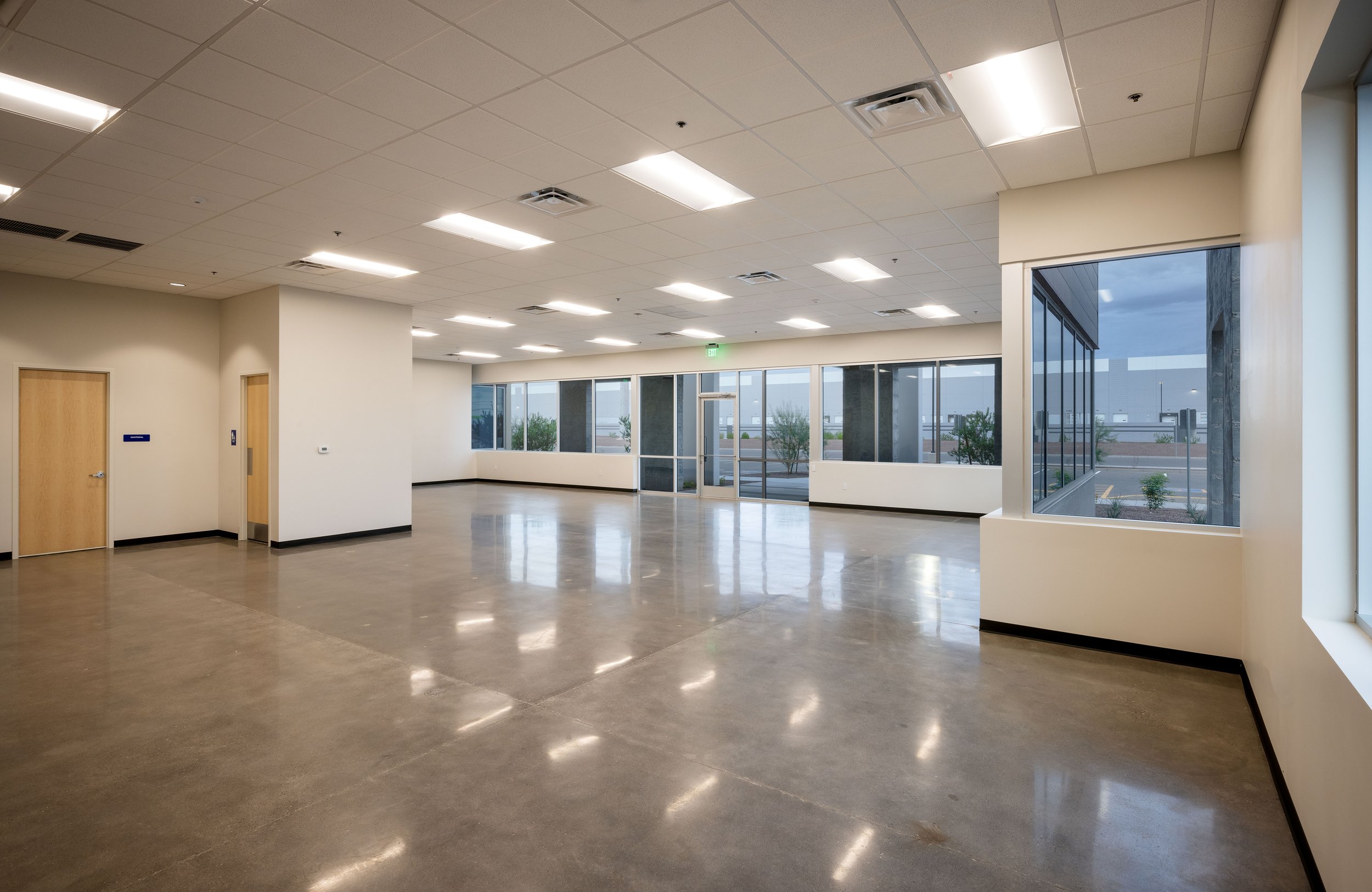 Empty commercial space with large windows, polished concrete floors, white walls, and a drop ceiling, with exterior view of a parking lot and cloudy sky.