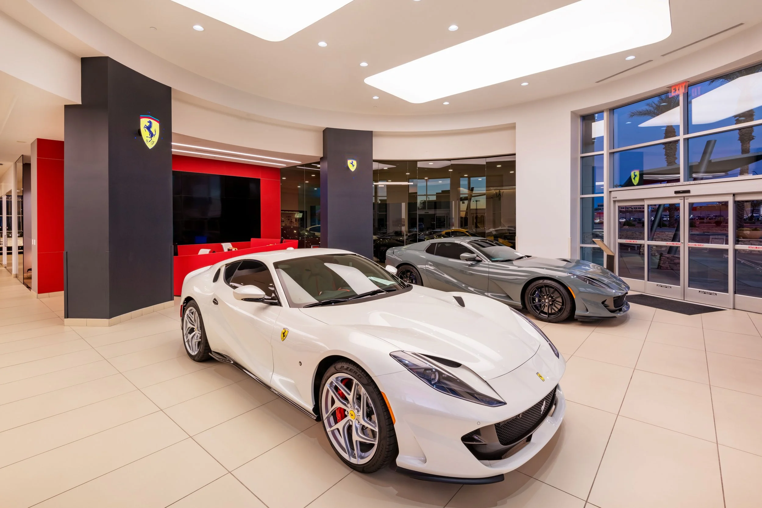 Inside a Ferrari dealership showing three luxury sports cars: a white Ferrari with black accents in the foreground, a silver Ferrari, and a dark gray Ferrari behind them, with large windows, modern ceiling lighting, and Ferrari logos on the pillars.
