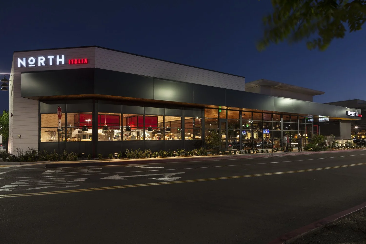 Night view of a modern restaurant building with large glass windows, illuminated inside, with the sign 'NORTH ITALIA' on the corner.