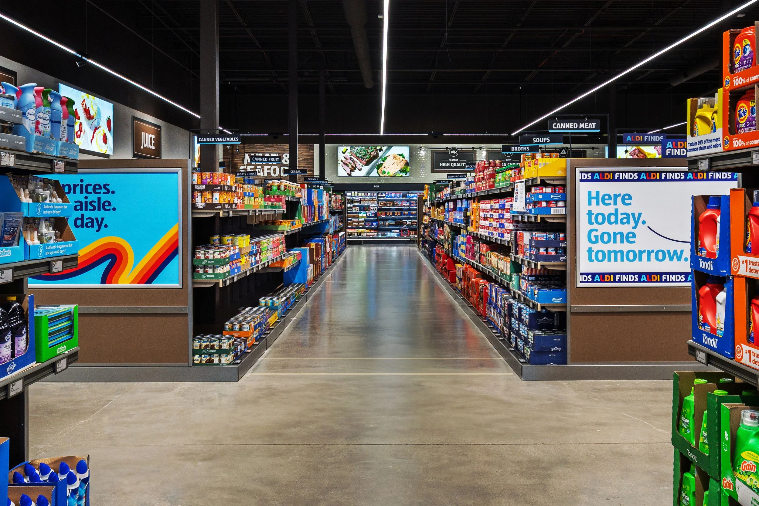 A grocery store aisle with shelves stocked with various food and household products, illuminated by overhead lighting, with digital displays on either side of the aisle.
