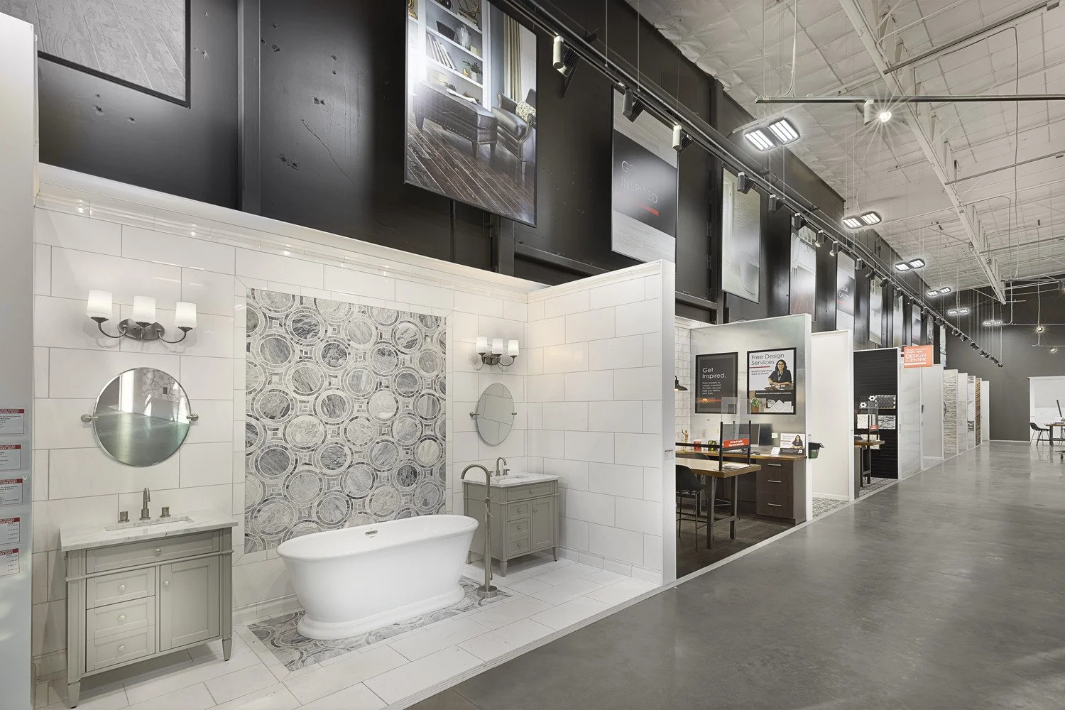 Showroom display of a bathroom with a freestanding bathtub, two vanities, mirrors, and wall-mounted sconces. Various interior design displays are visible in the background.