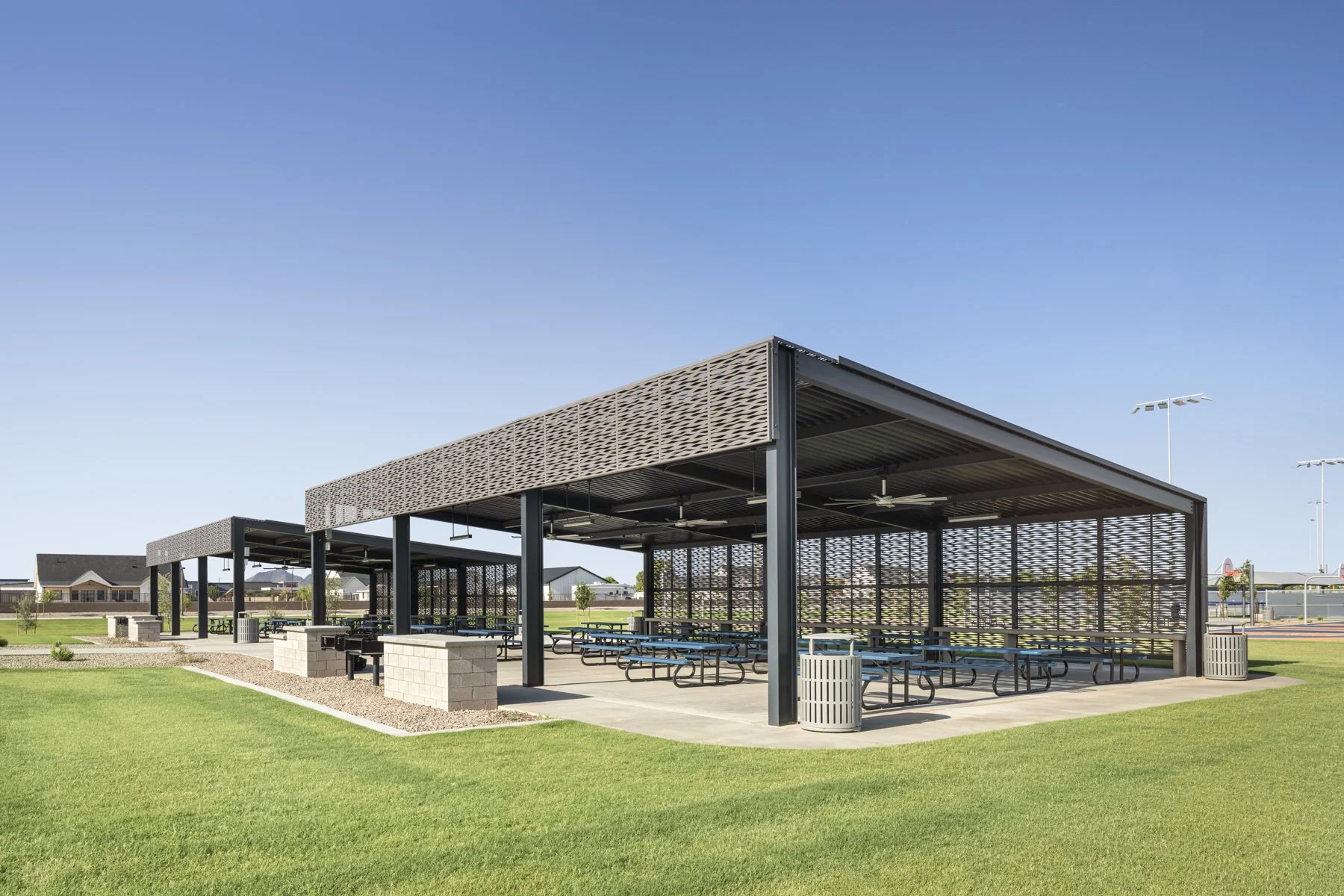 A covered outdoor picnic area with metal benches, surrounded by green grass and a few trees, under a clear blue sky.