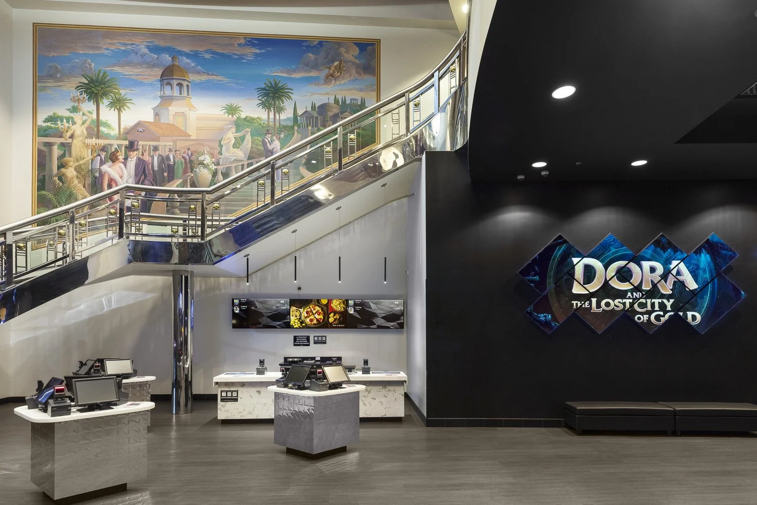 Interior of a theater lobby with a large TV screen displaying the title "Dora and the Lost City of Gold" on a black wall, a staircase with a glass and metal railing leading to an upper level, a large colorful mural with palm trees and people in forma