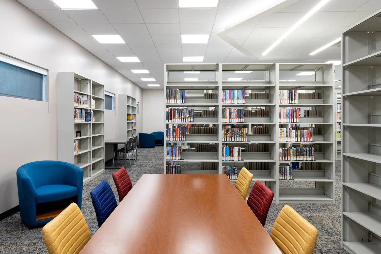 Interior of a modern library with bookshelves, a wooden table with colorful chairs, and a cozy seating area with blue chairs. Shelves contain many books, and the room has white walls and ceiling with bright lighting.