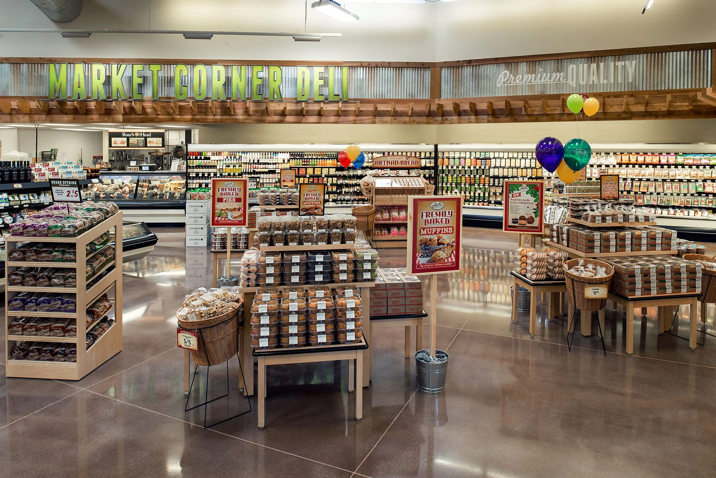 Interior of a grocery store with displays of baked goods like muffins and pies, and balloons for promotion, under a sign that reads 'Market Corner Deli'.