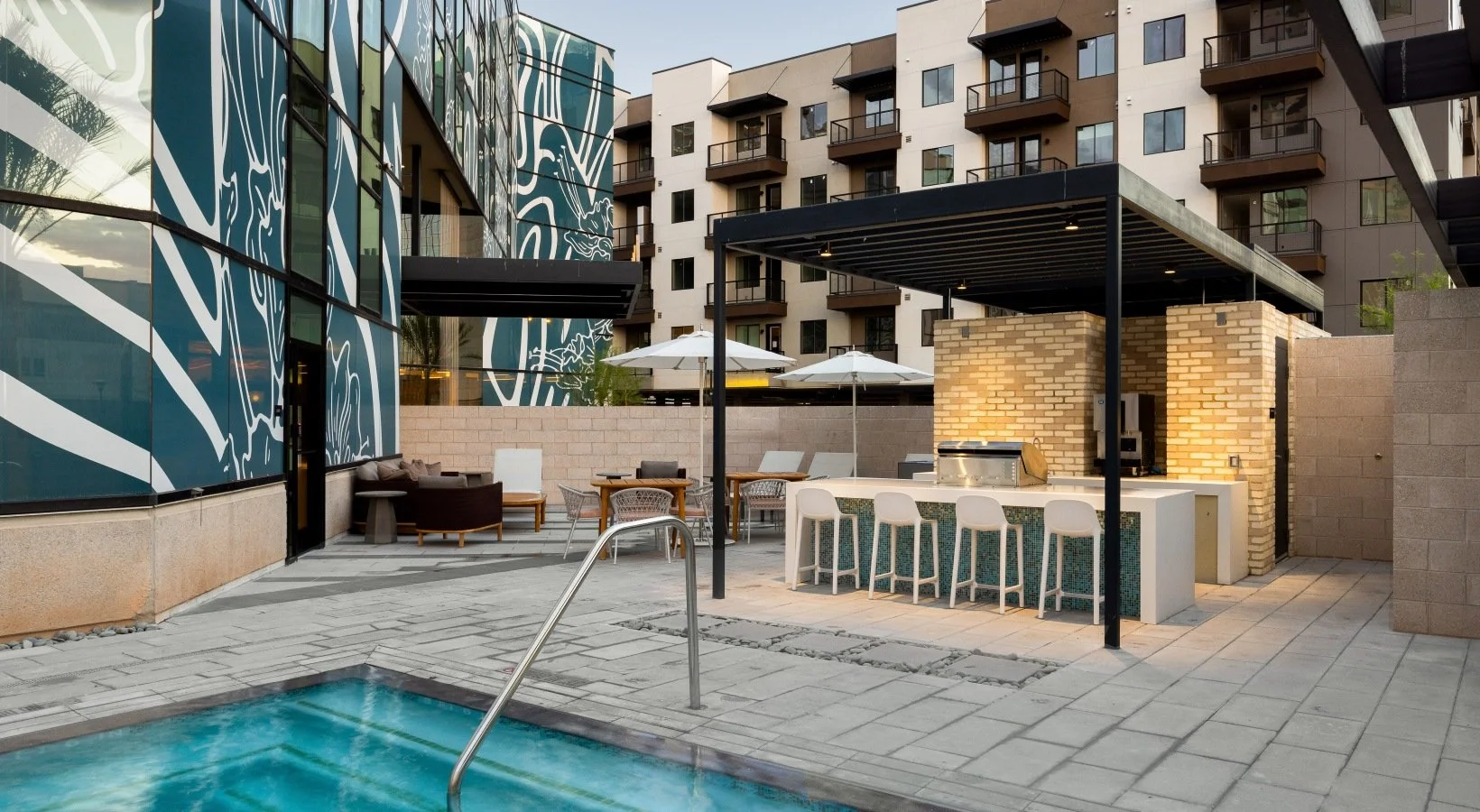 Outdoor pool area with seating, umbrellas, and a bar with barstools under a black roof, surrounded by modern apartment buildings.