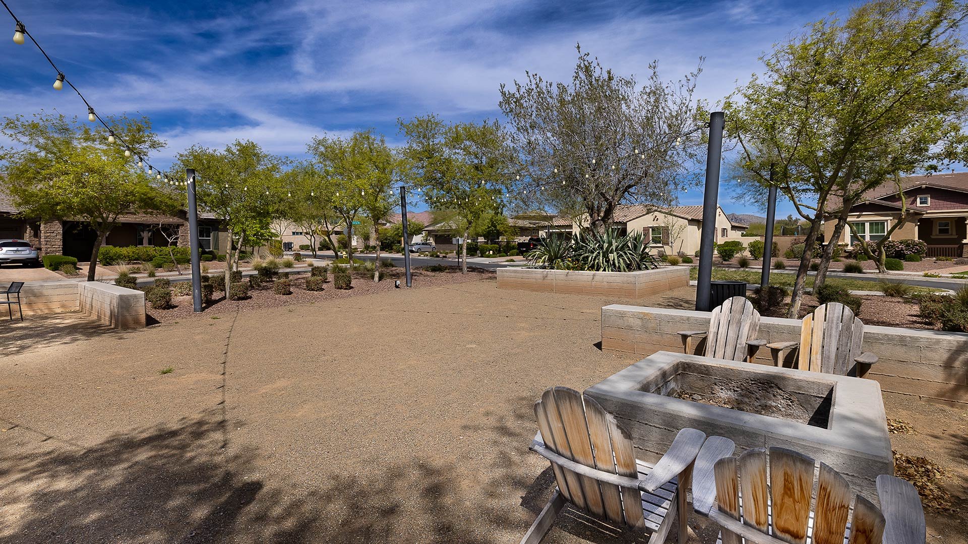 Quiet neighborhood park with trees, string lights, and wooden chairs around a fire pit under a blue sky with scattered clouds.