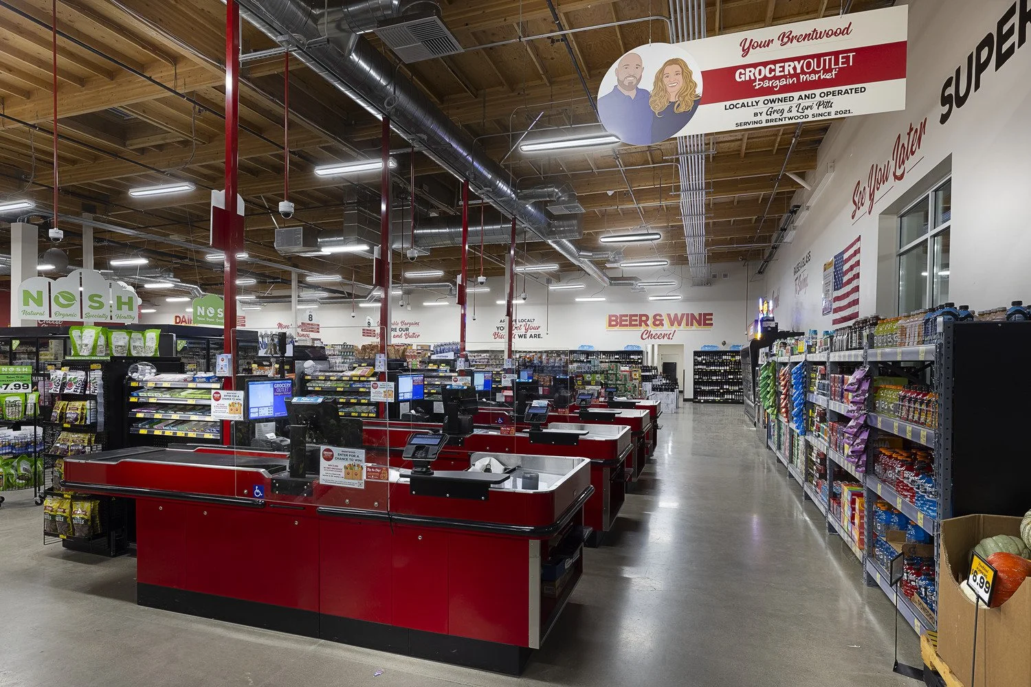 Interior view of a grocery store checkout area with multiple cash registers, red counters, and a variety of grocery shelves on the sides. Signage indicates sections for beer, wine, and a grocery outlet. Bright lighting and wooden ceiling with visible