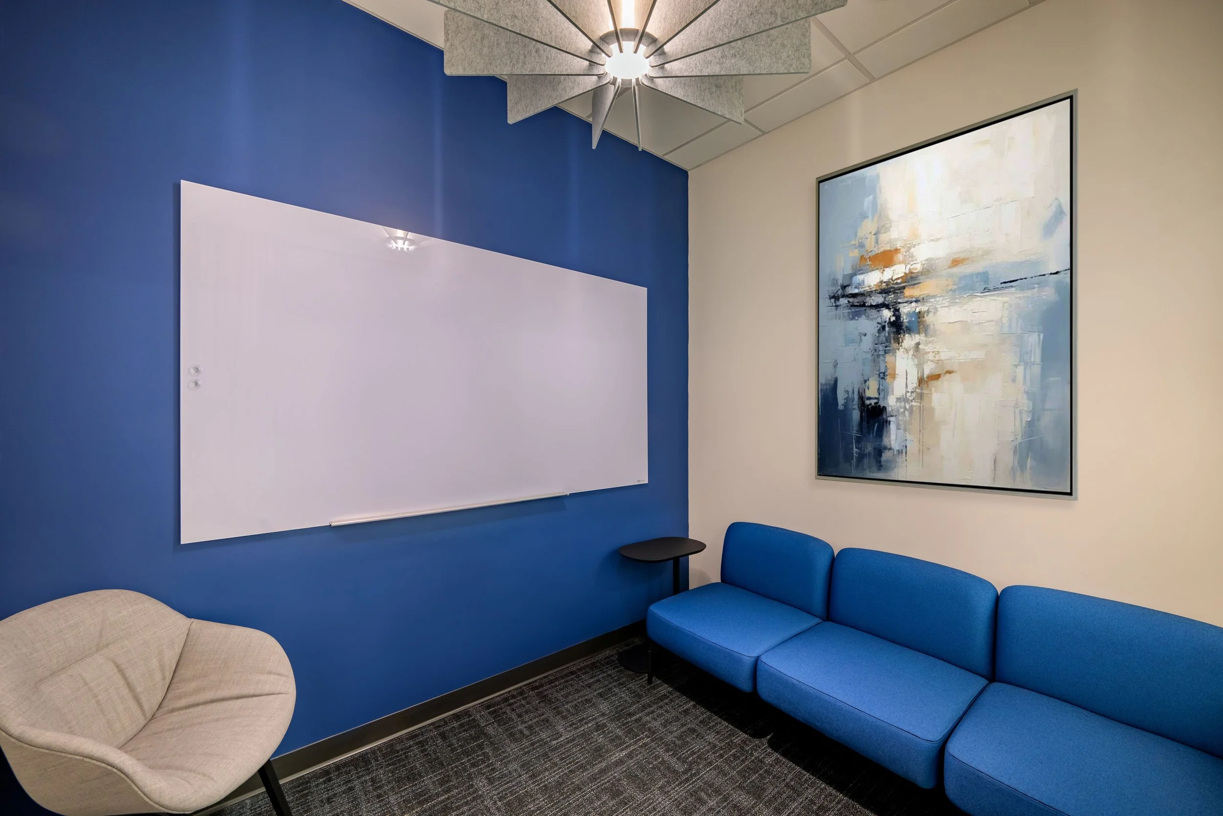Office waiting room with blue wall, abstract painting, blue sofa, beige chair, small side table, whiteboard, and ceiling fan.