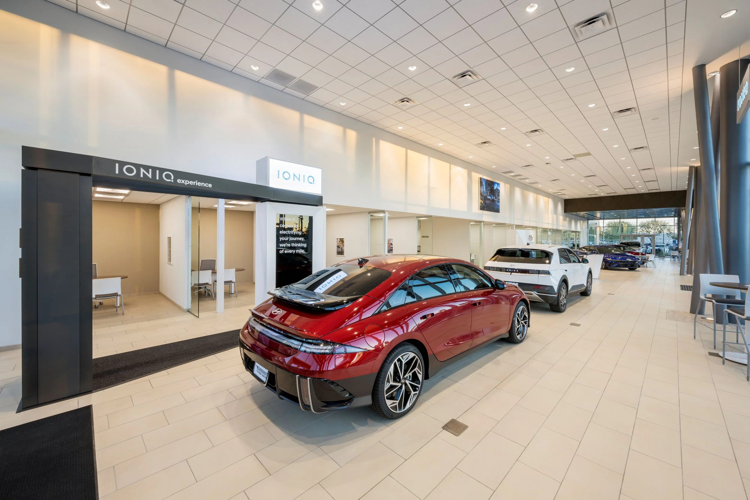 Inside a car dealership showroom with multiple electric vehicles on display, including a red car in the foreground, and white and dark-colored cars parked further back. The space has large windows, modern design, and seating areas.
