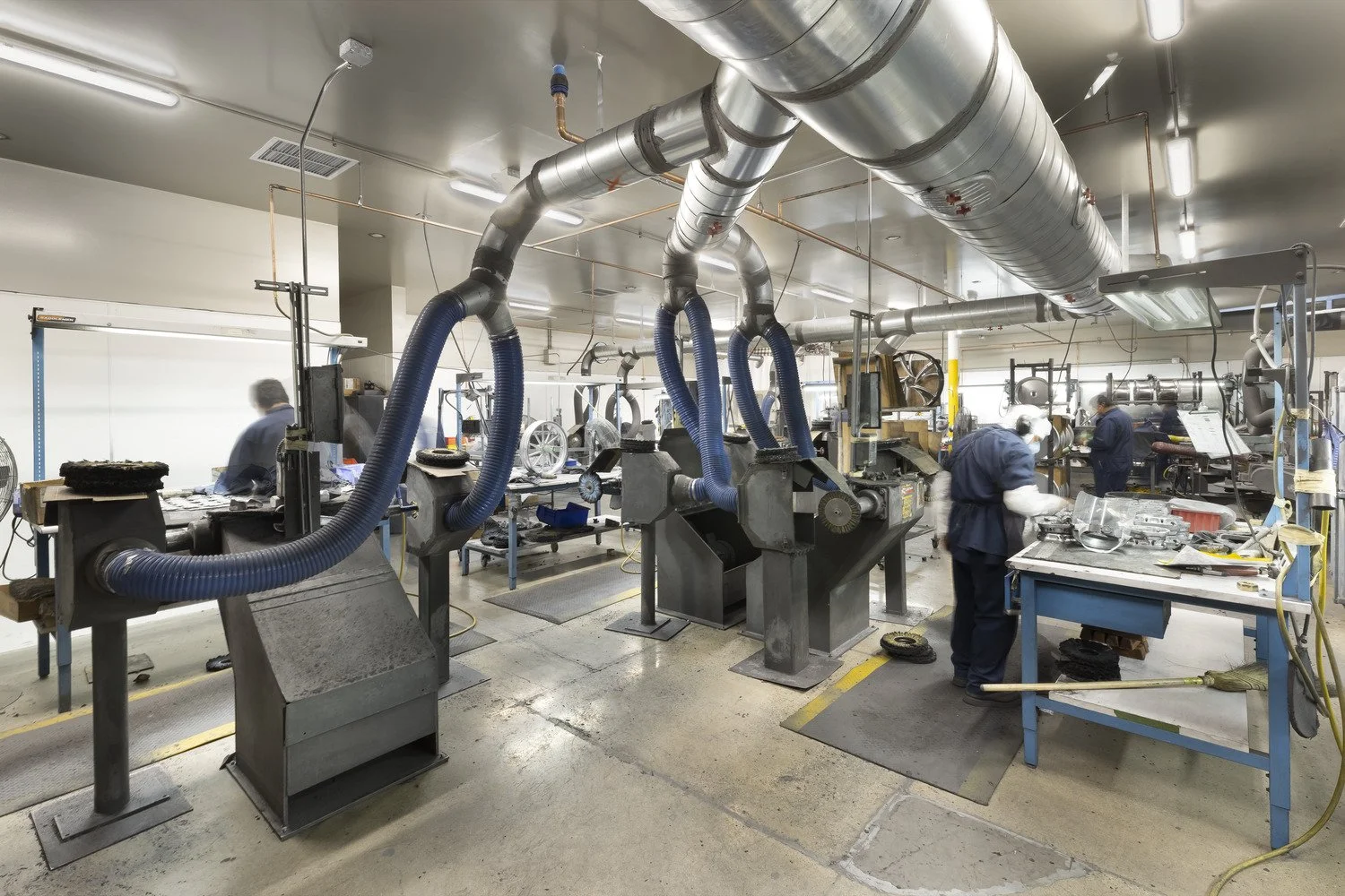 Workers in a manufacturing factory with machinery, large ventilation pipes, and workbenches.