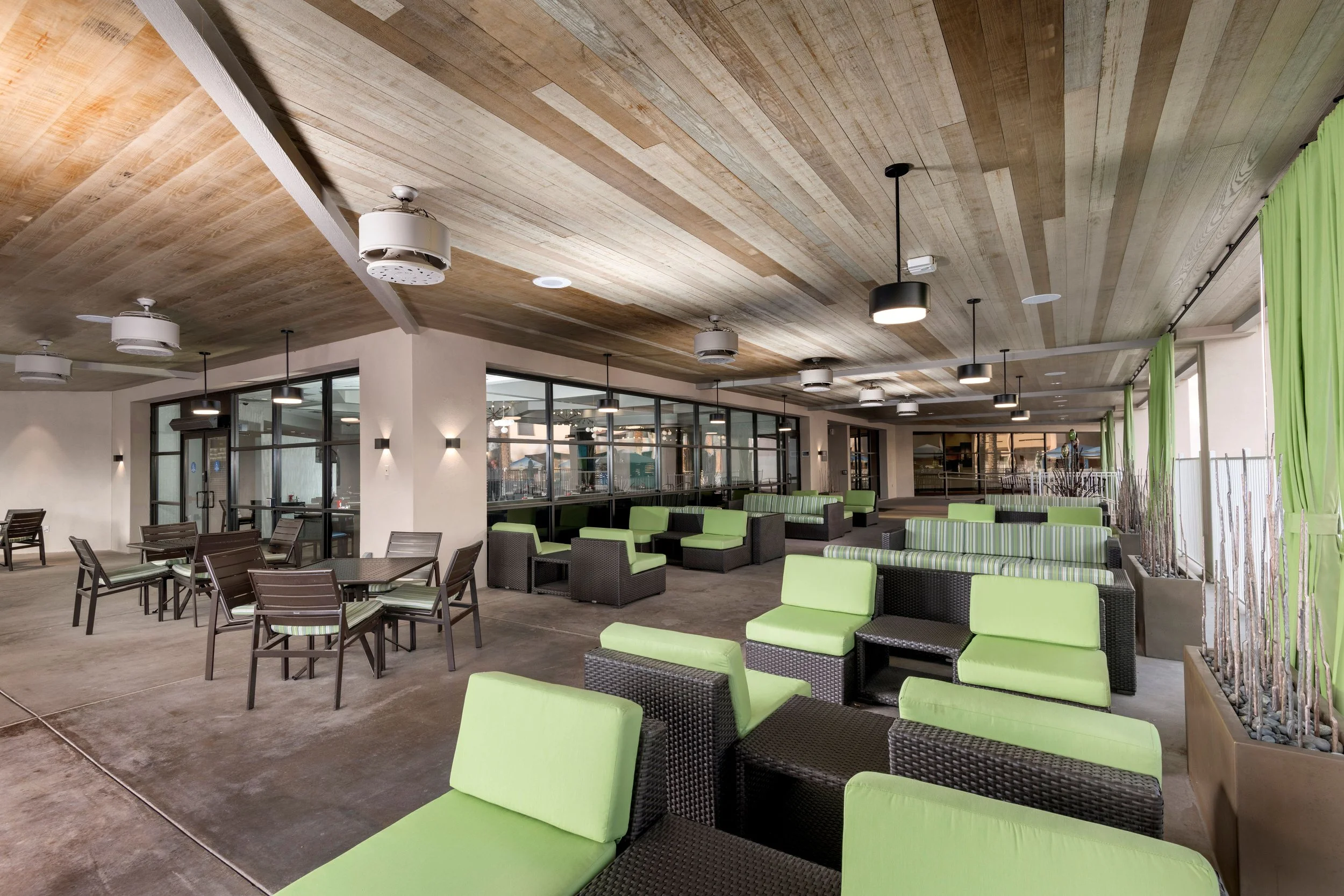 Indoor lounge area with black wicker chairs and sofas with light green cushions, large windows with green curtains, and a wooden ceiling.