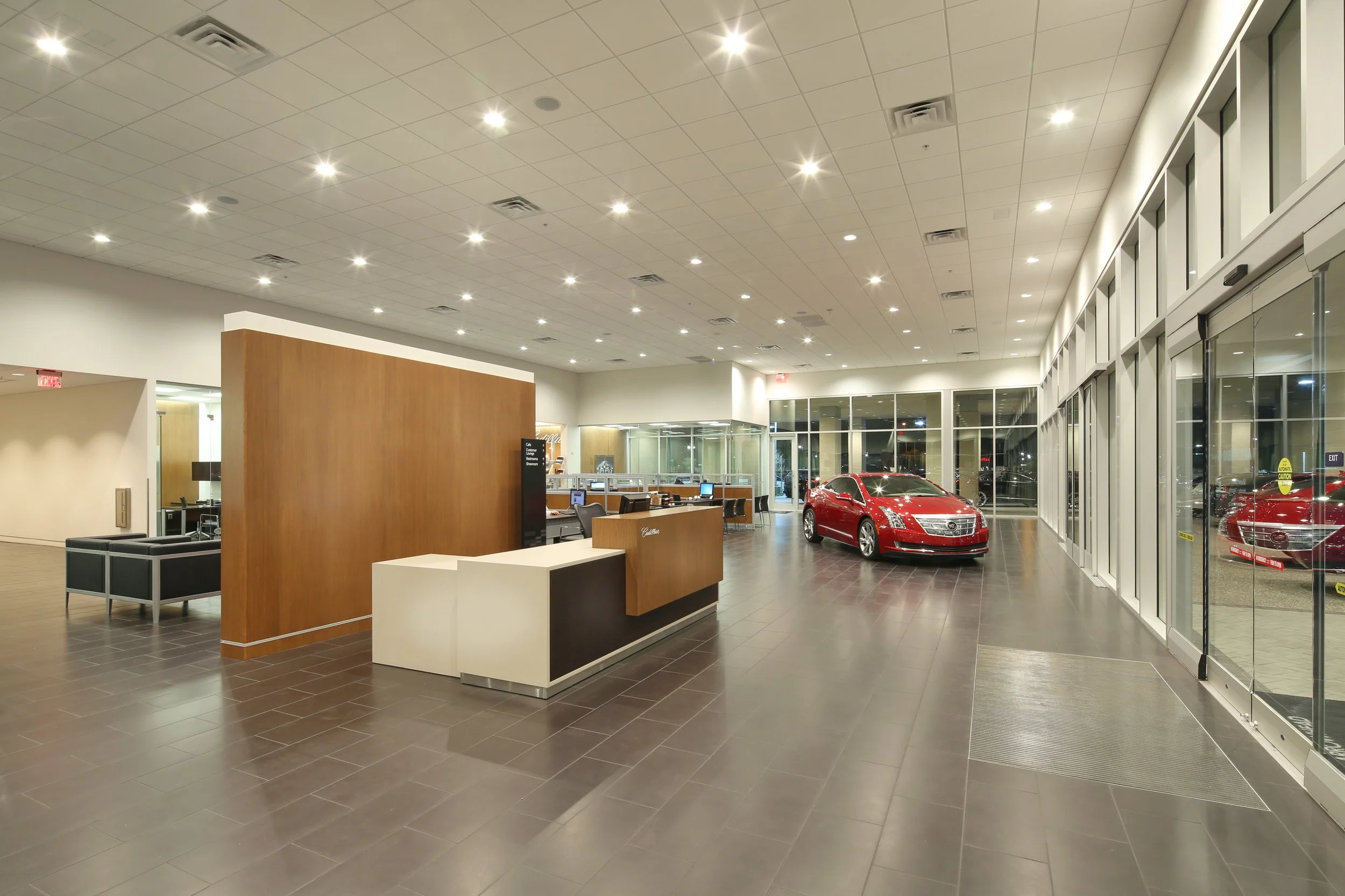 A car showroom with a red luxury car near the glass windows and a reception desk, with seating and computers in the background.
