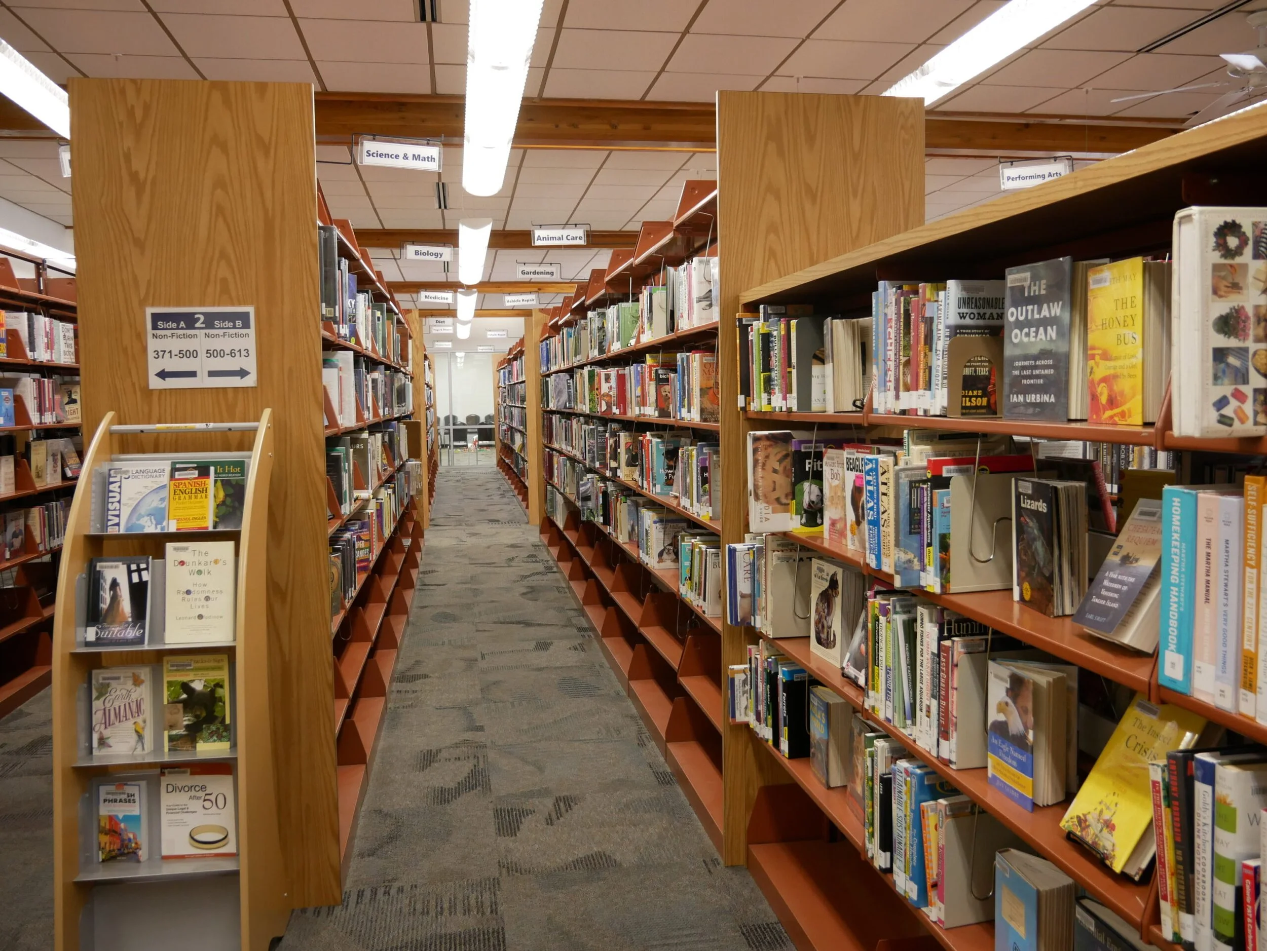 A view of bookshelves in a library aisle, with signs indicating sections like Science & Math, Biology, Animal Care, Gardening, Performing Arts. The aisle is carpeted and well-lit.