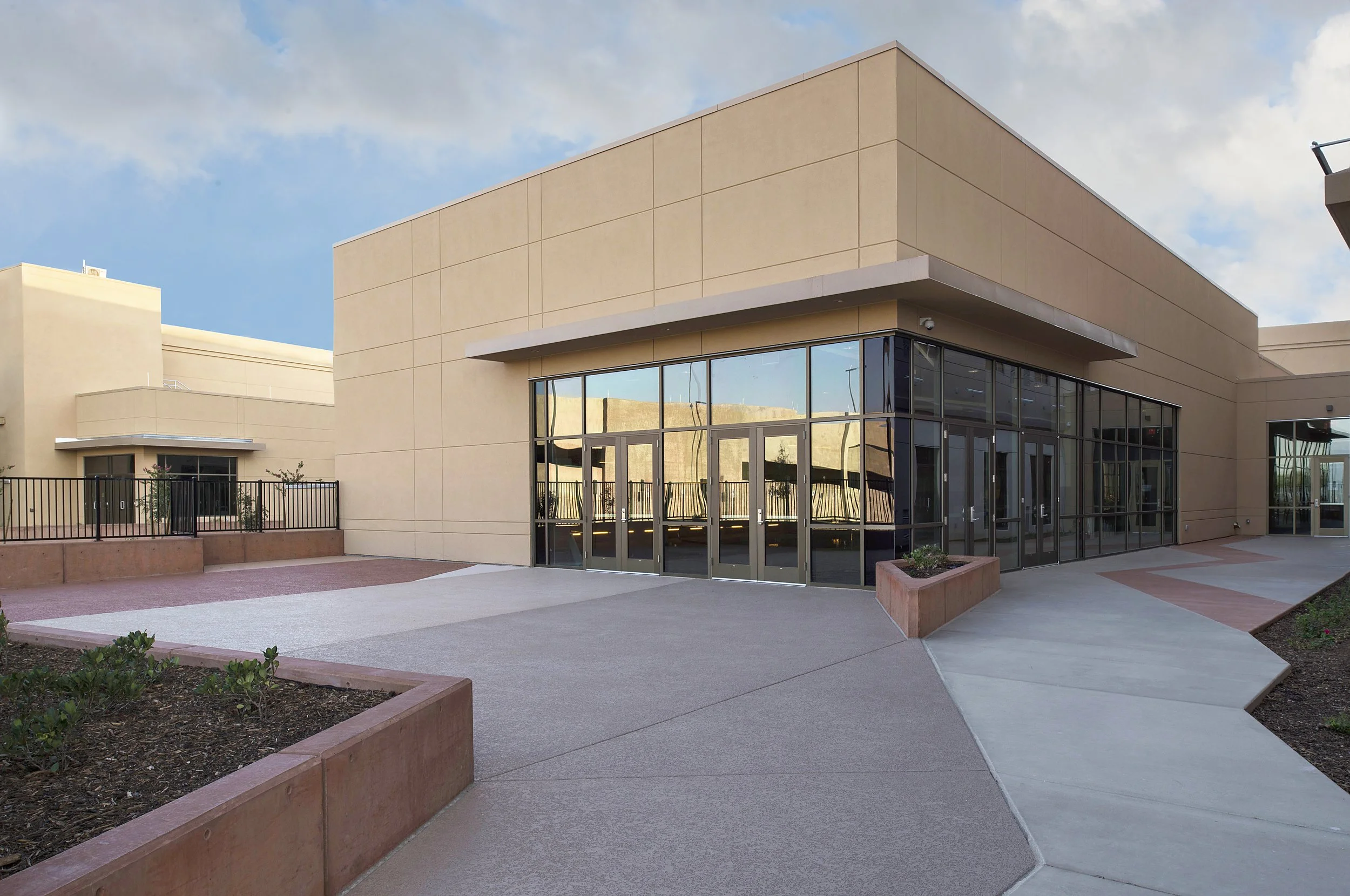 Modern building with large glass windows and beige walls, surrounded by paved walkways and landscaped areas with small plants.