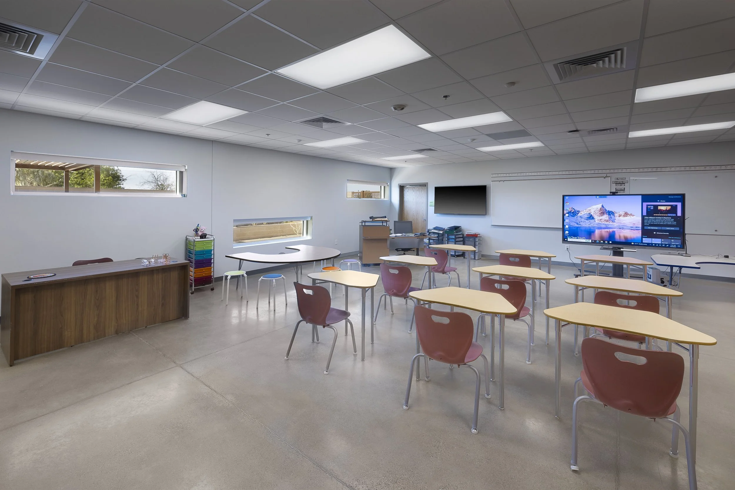 Empty classroom with yellow desks and pink chairs, two windows, a wooden teacher's desk, a toy storage cart, a large screen at the front, and a whiteboard.