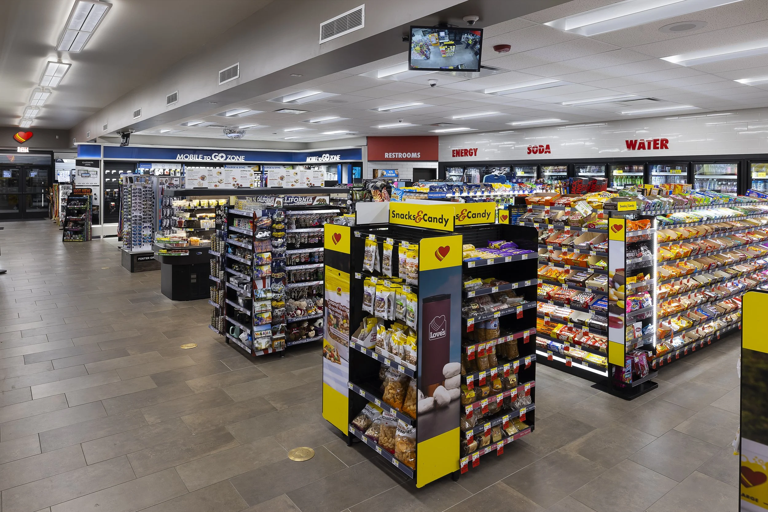 Interior of a convenience store with shelves of snacks, candy, soda, water, and other products, with signs for restrooms, energy, soda, and water at the back.
