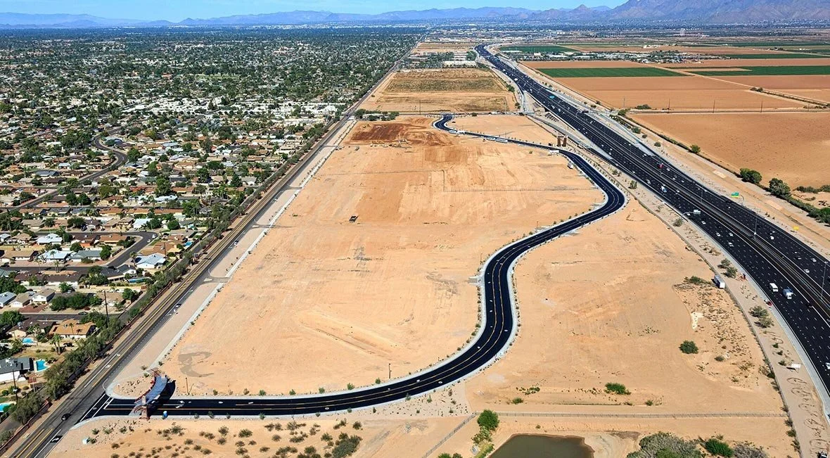 An aerial view of a highway under construction next to a residential neighborhood and surrounding farmland under a clear blue sky.