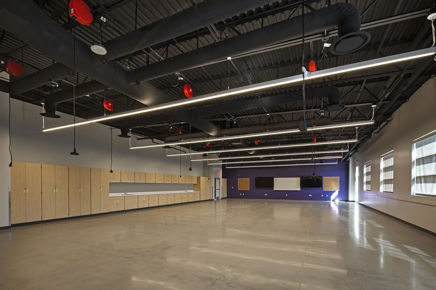 Empty modern classroom or training room with polished concrete floor, wooden cabinets along the left wall, purple accent wall with mounted TV screens, large windows with striped blinds, black ceiling with exposed ductwork, hanging linear LED lights, 